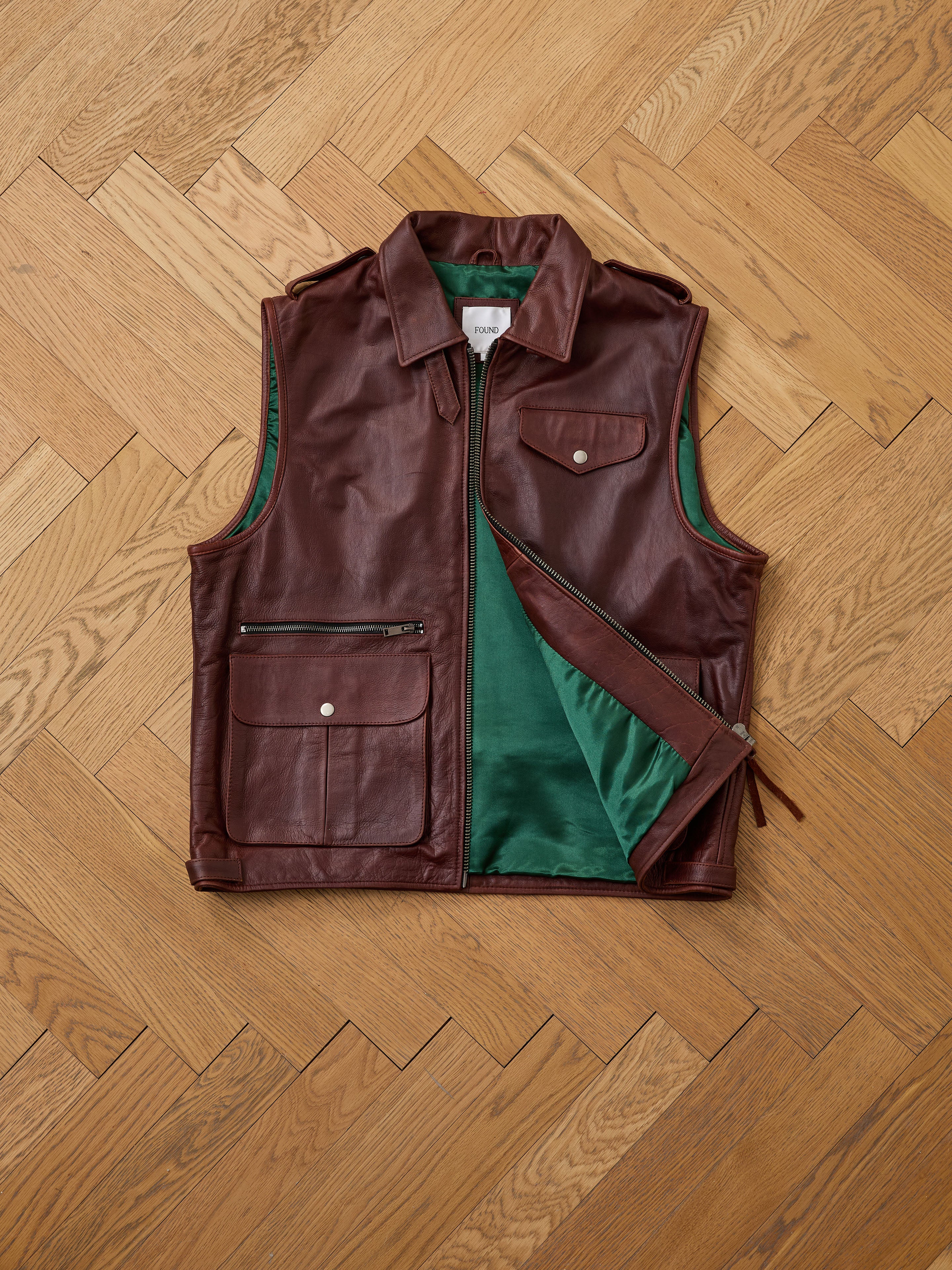 The Found Leather Pocket Vest, in distressed brown leather with utility pockets and a zipper, is shown open to display its green lining, laid flat on a wooden herringbone floor.