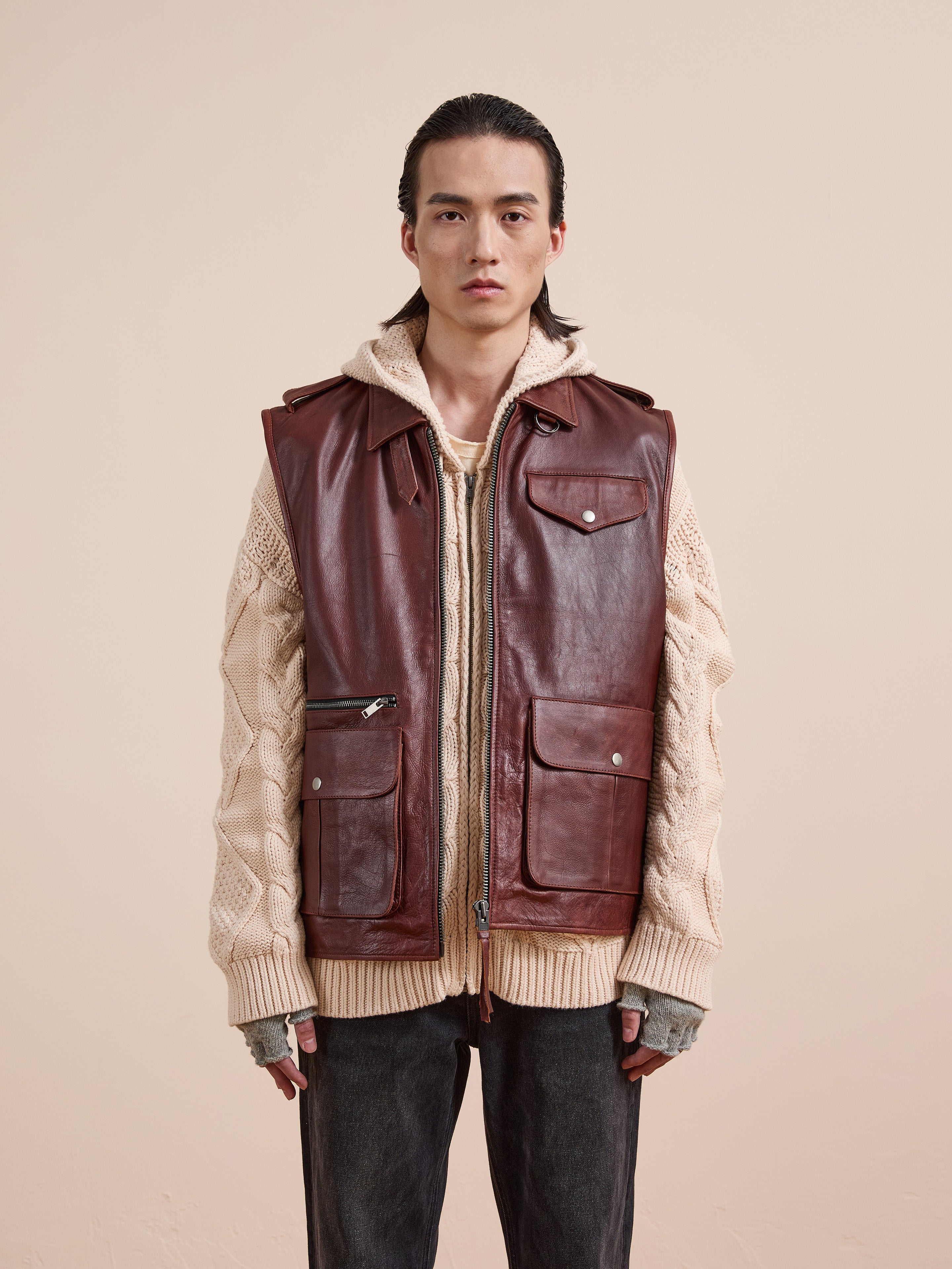 A person wears a cream cable-knit sweater layered under the Found Leather Pocket Vest in distressed brown, paired with dark pants against a plain beige background.