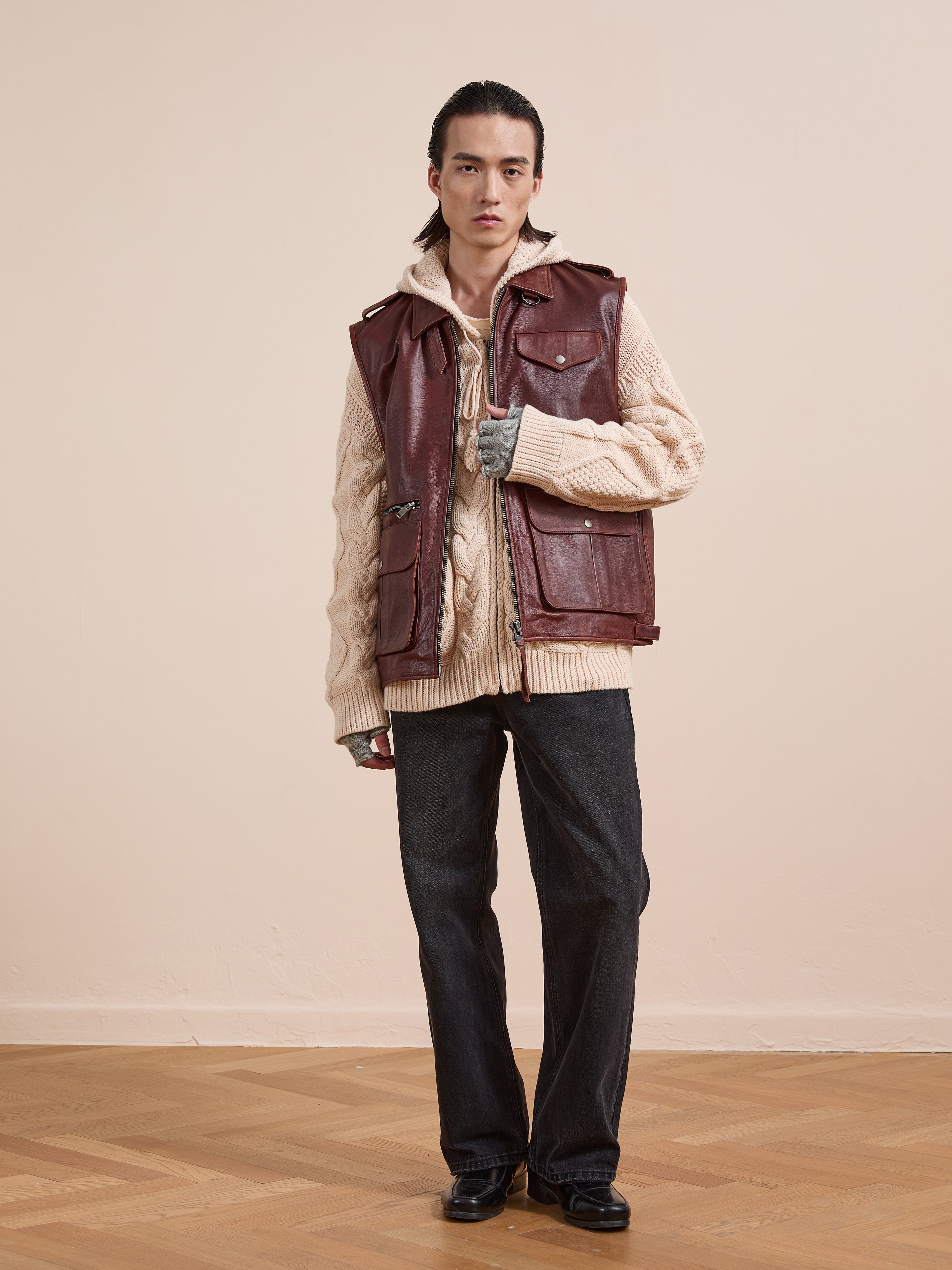 A person stands indoors wearing Found’s maroon Leather Pocket Vest with utility pockets over a beige cable-knit sweater, dark wide-leg pants, black shoes, and light gray fingerless gloves.