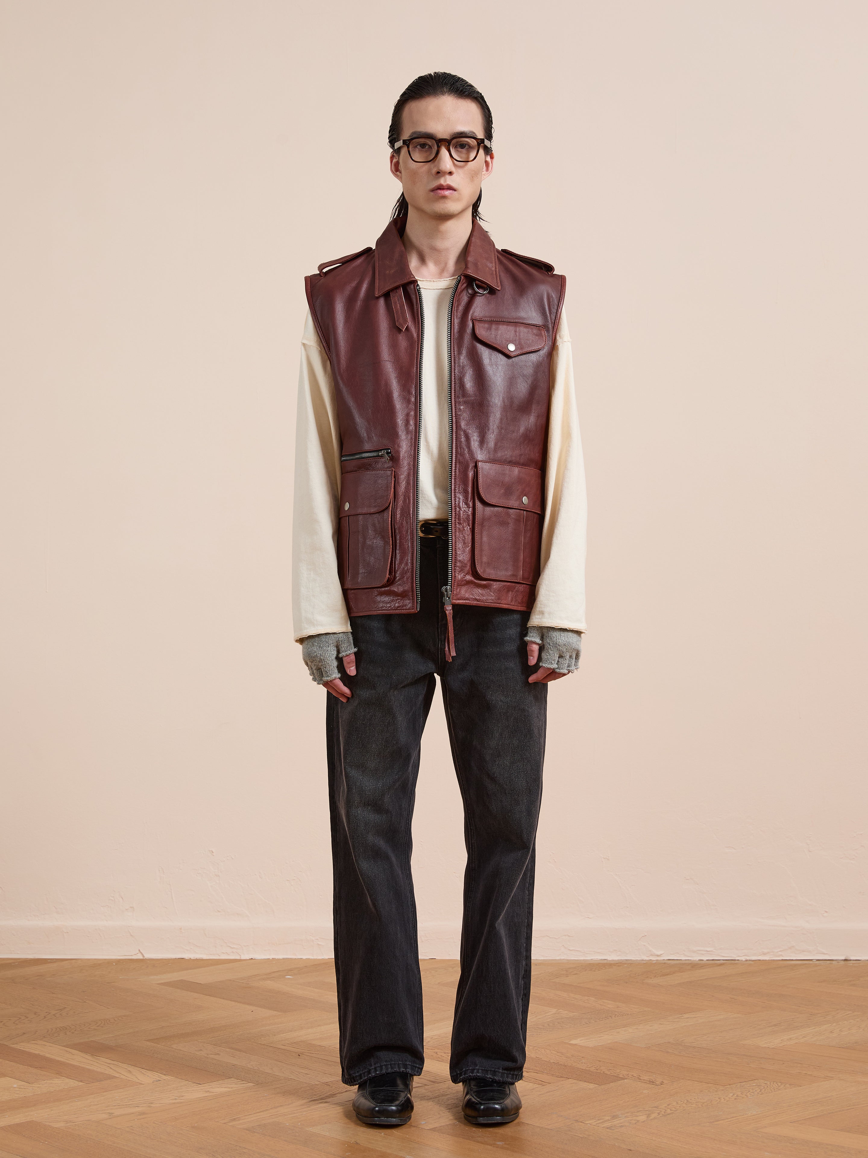 A person stands indoors on a wooden floor, facing forward, wearing the Found Leather Pocket Vest in maroon with utility pockets over a cream long-sleeve shirt, dark jeans, black shoes, glasses, and fingerless gloves.