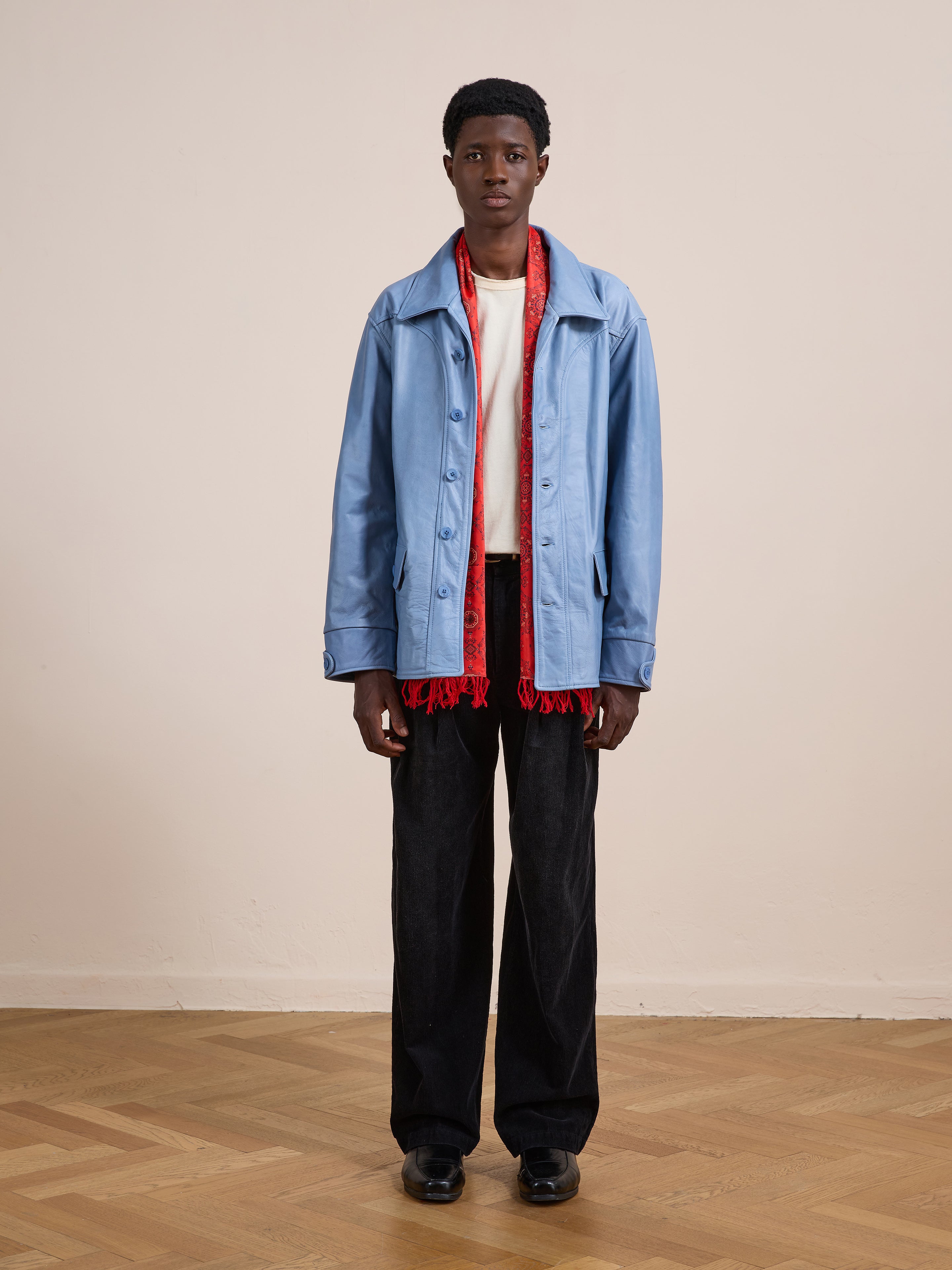 A person stands indoors in a relaxed fit FOUND Leather Field Jacket, white shirt, black pants, black shoes, and a red scarf against a plain beige wall and wooden floor.