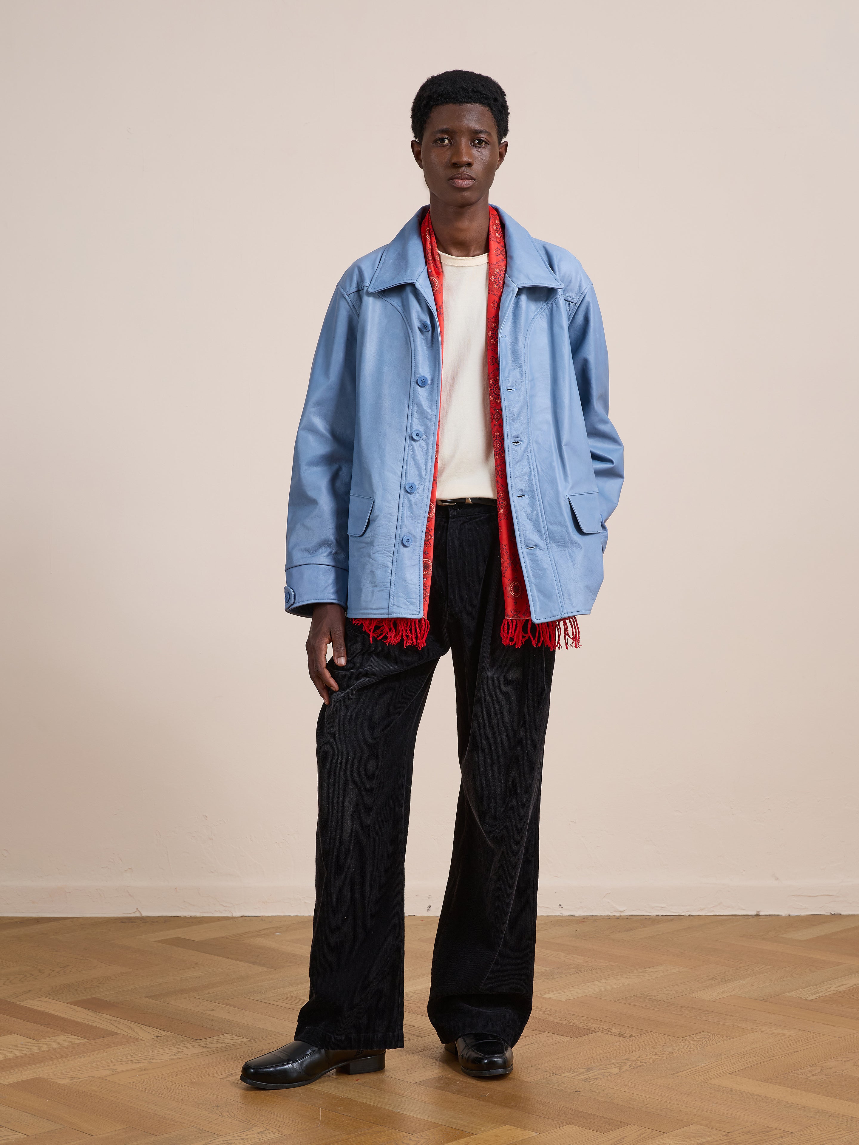 A person stands indoors on a wooden floor and beige wall, wearing the FOUND Leather Field Jacket in a relaxed fit, white shirt, red scarf, unisex black wide-leg pants, and black shoes.