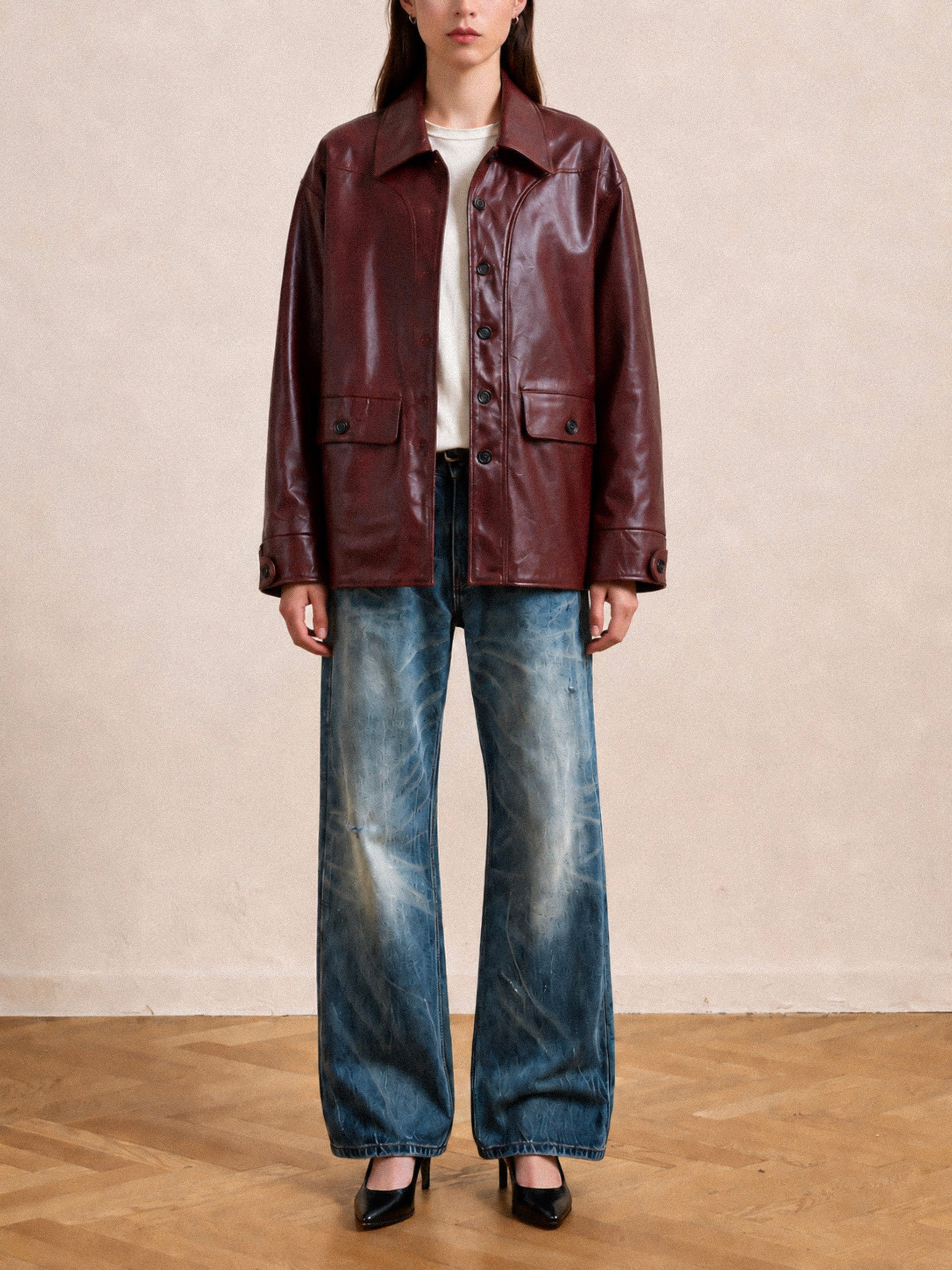 A person stands on a wooden floor against a plain light background, wearing the FOUND Leather Field Jacket with a relaxed fit over a white shirt, paired with faded blue wide-leg jeans and black heels.