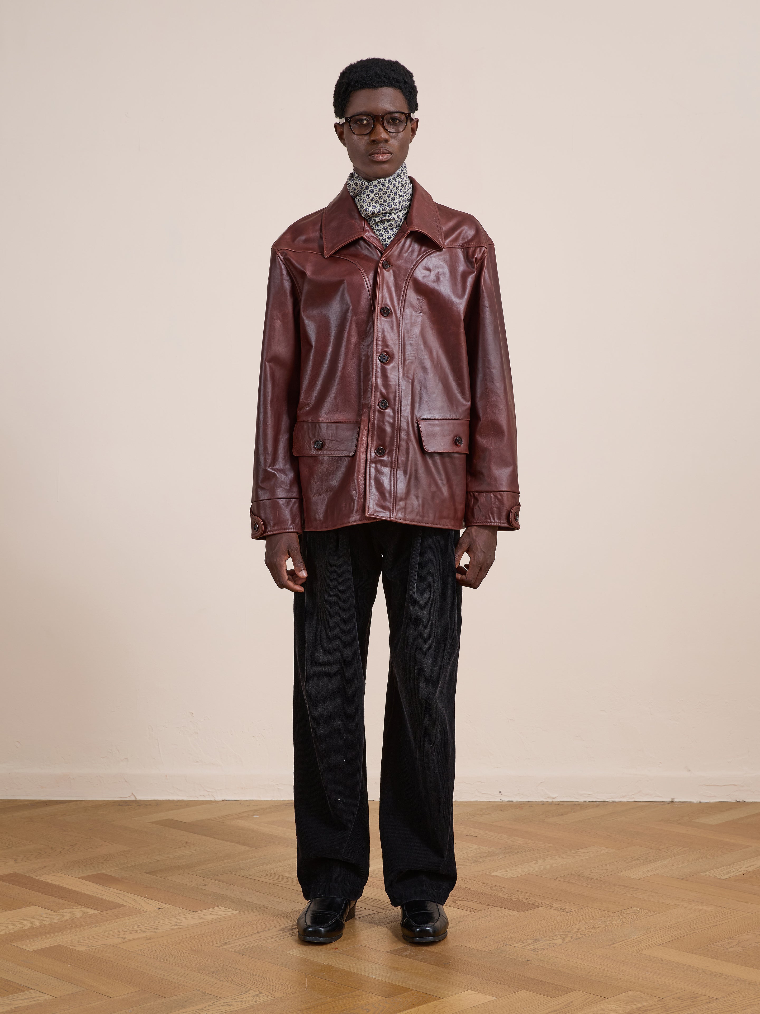 A person stands indoors wearing a relaxed fit FOUND Leather Field Jacket, patterned turtleneck, black pants, and black shoes against a plain beige wall and wooden parquet floor.