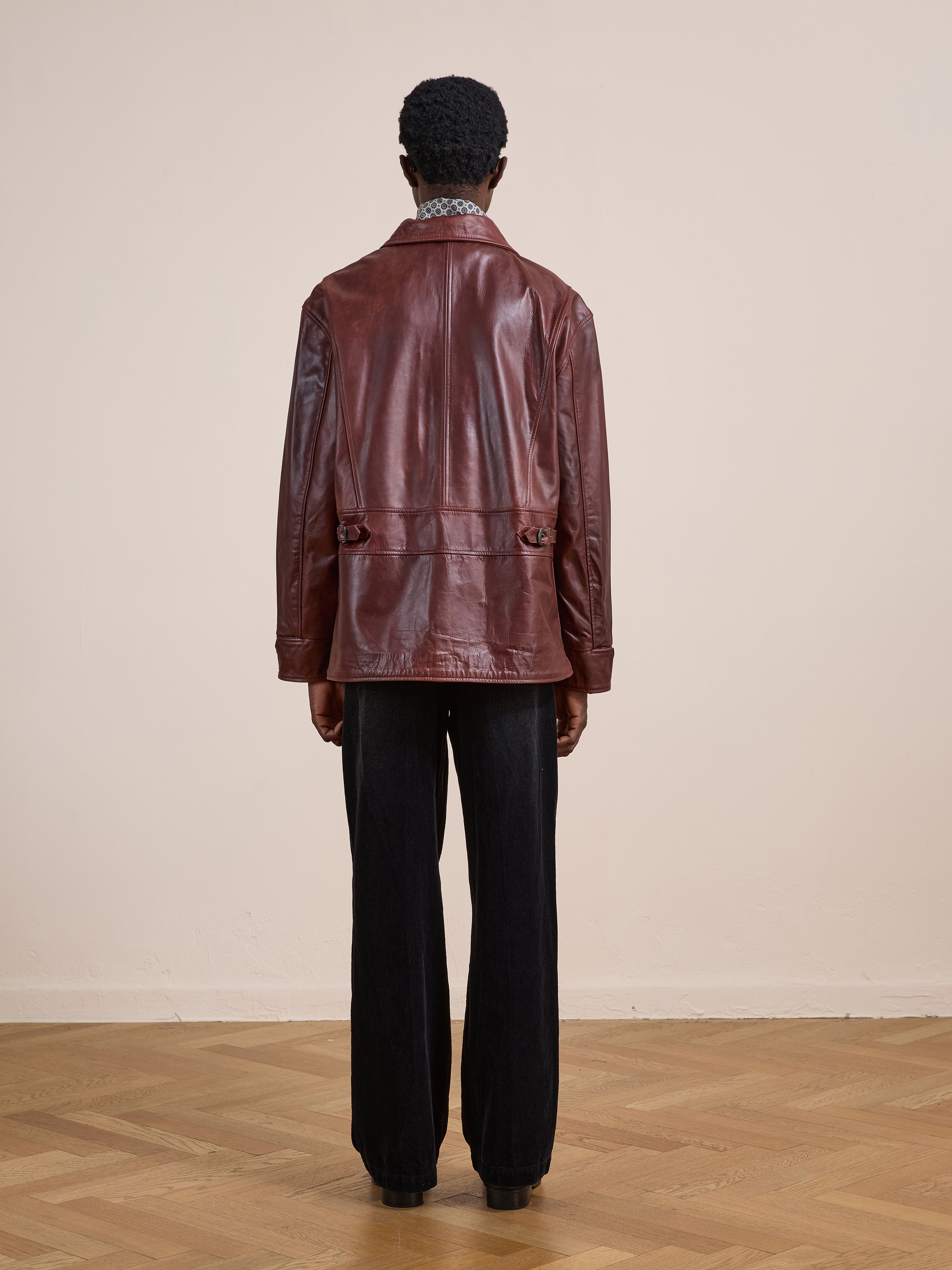 A person stands indoors on a wooden floor, facing away from the camera, wearing the FOUND Leather Field Jacket in a relaxed fit and black pants.