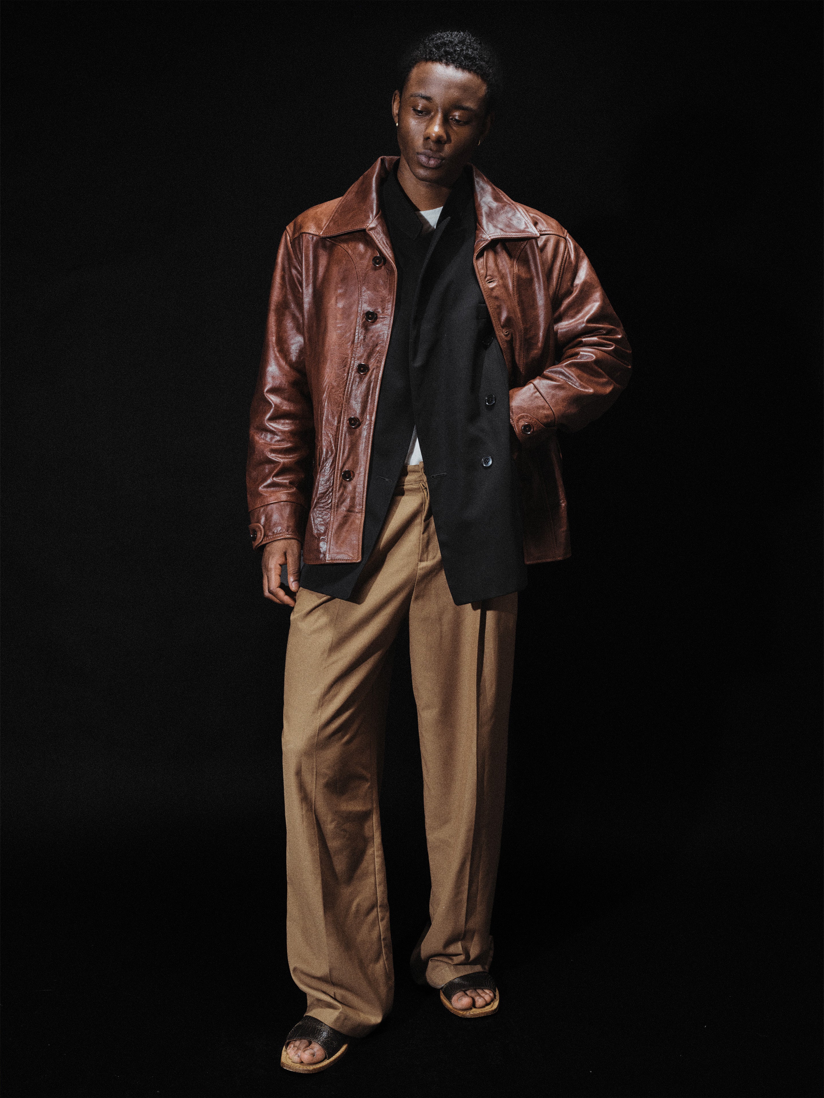 A man stands against a black background wearing the FOUND Leather Field Jacket in a classic workwear style, paired with a black shirt, tan wide-leg pants in a unisex relaxed fit, and sandals.