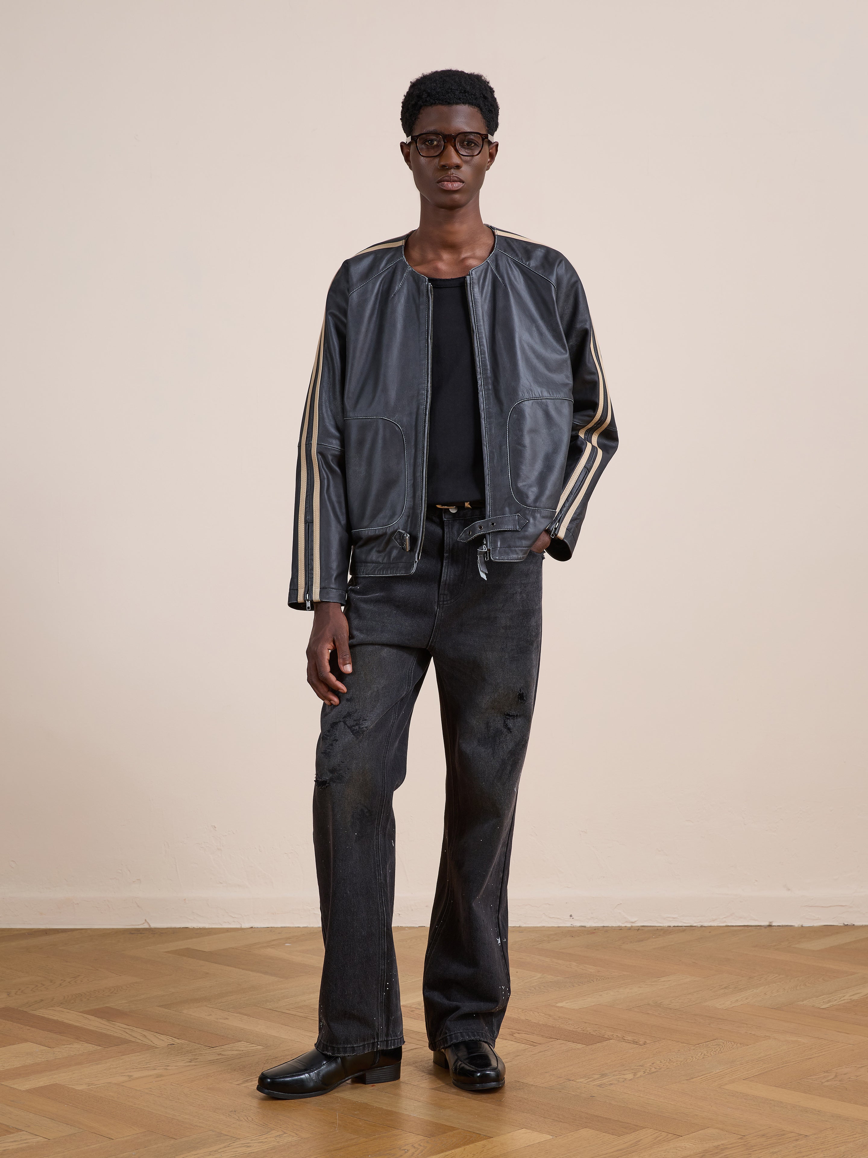 A person stands indoors against a beige wall and wooden floor, wearing the FOUND Leather Track Jacket with striped sleeves, paired with a black shirt, jeans, and shoes.
