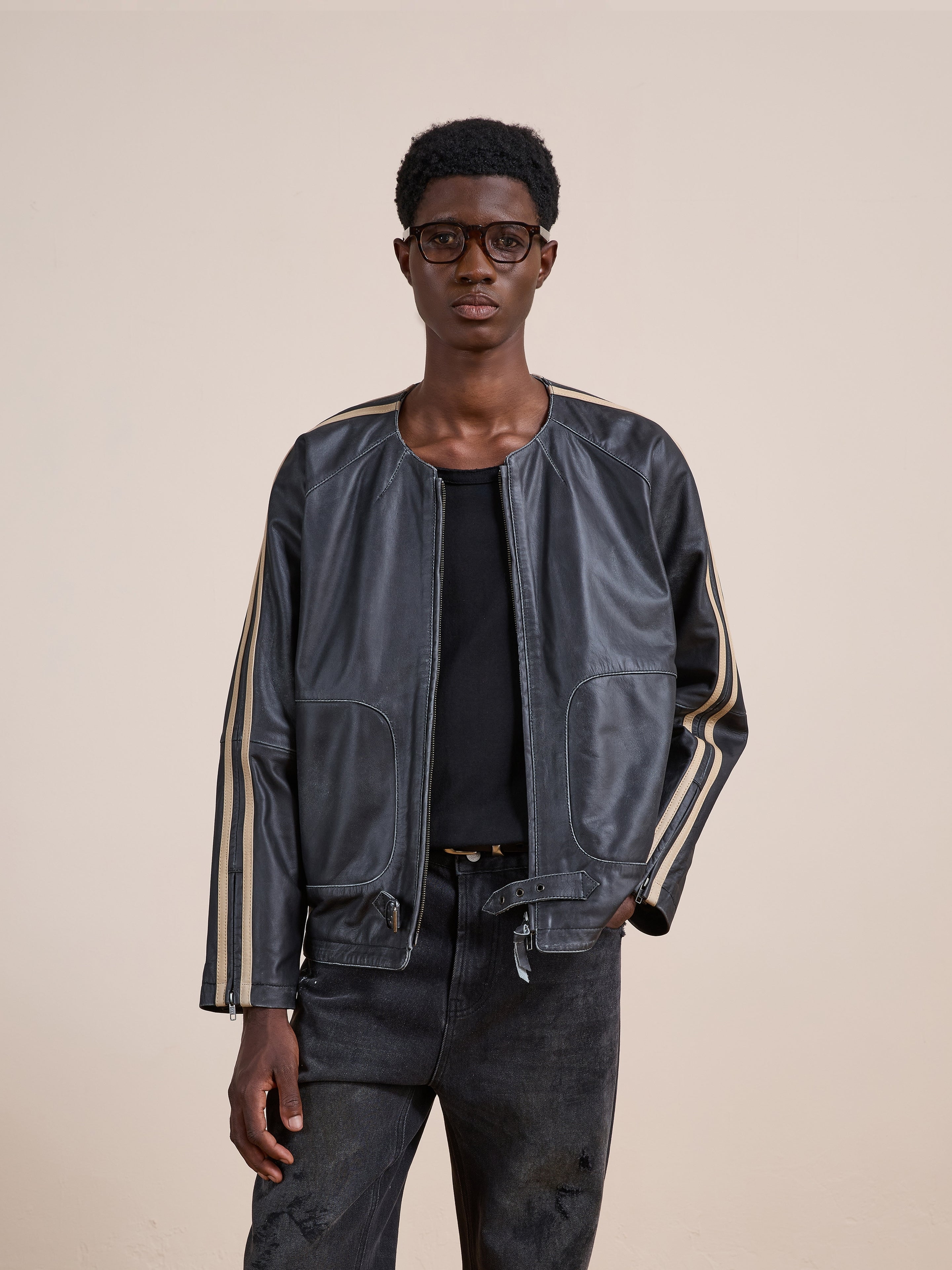 A man wearing glasses and a black shirt styles the FOUND Leather Track Jacket, featuring vintage-inspired beige stripes, while standing against a plain beige background.