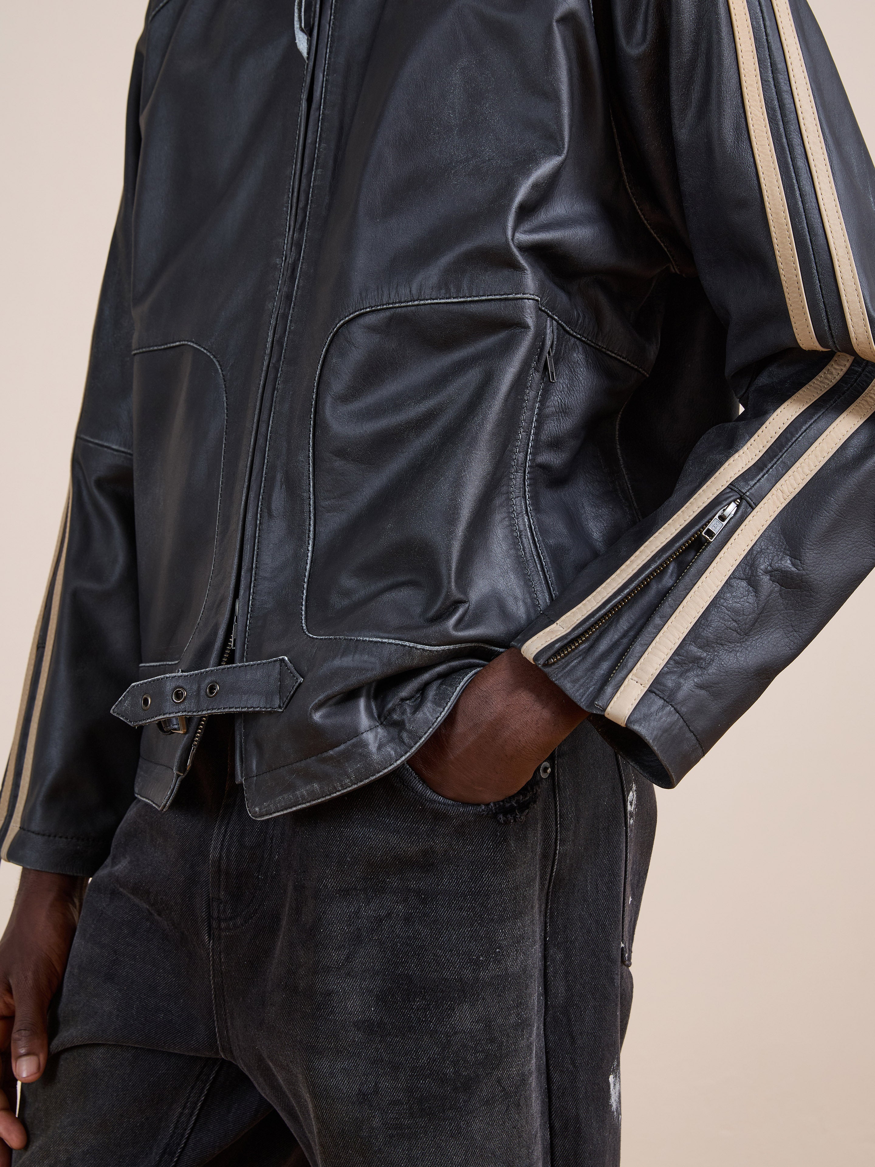 A person wearing the FOUND Leather Track Jacket with beige sleeve stripes and black jeans stands with one hand in their pocket.