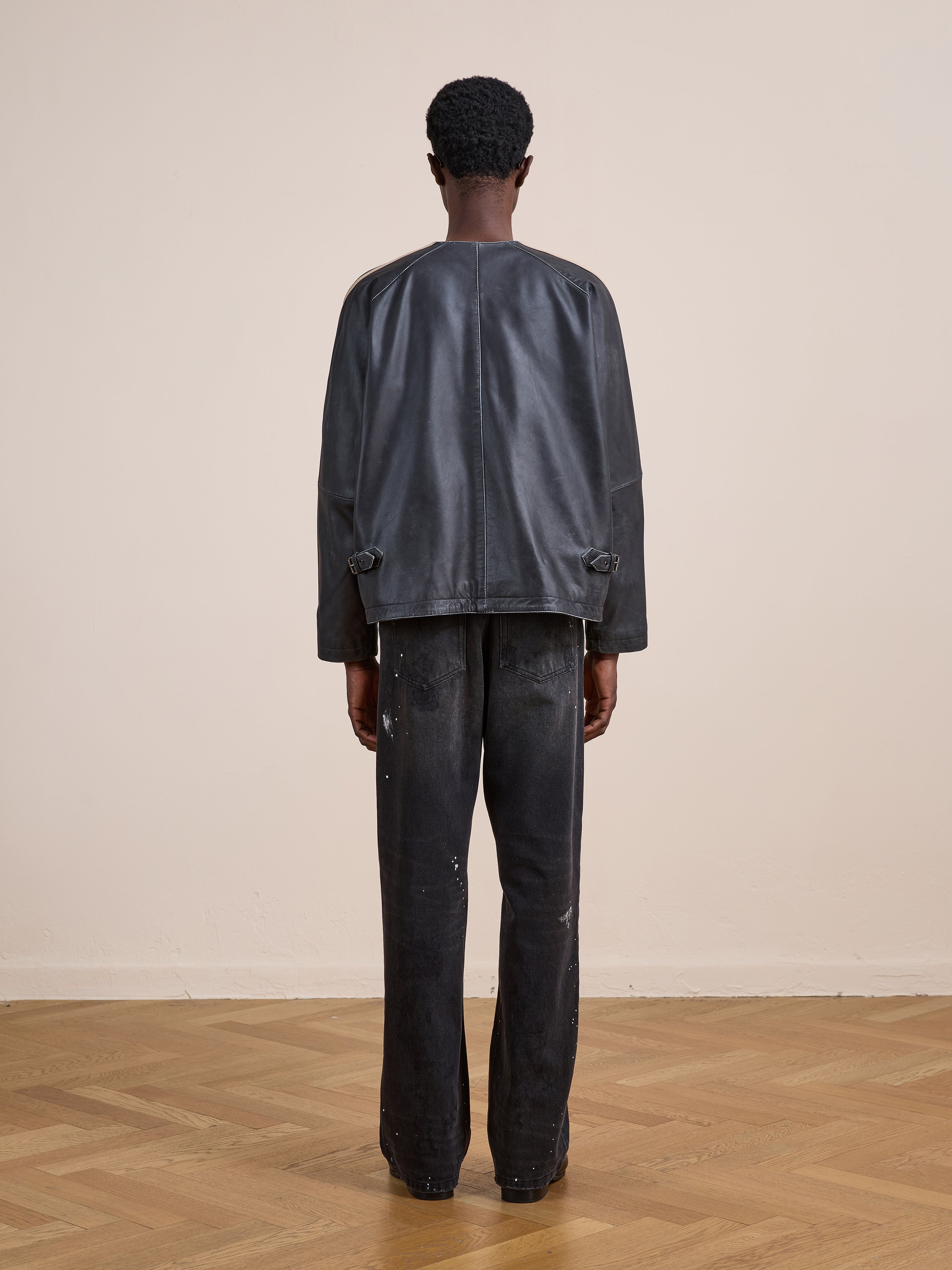 A person stands indoors on a wooden floor, facing away from the camera, wearing FOUND's Leather Track Jacket and black paint-splattered pants.