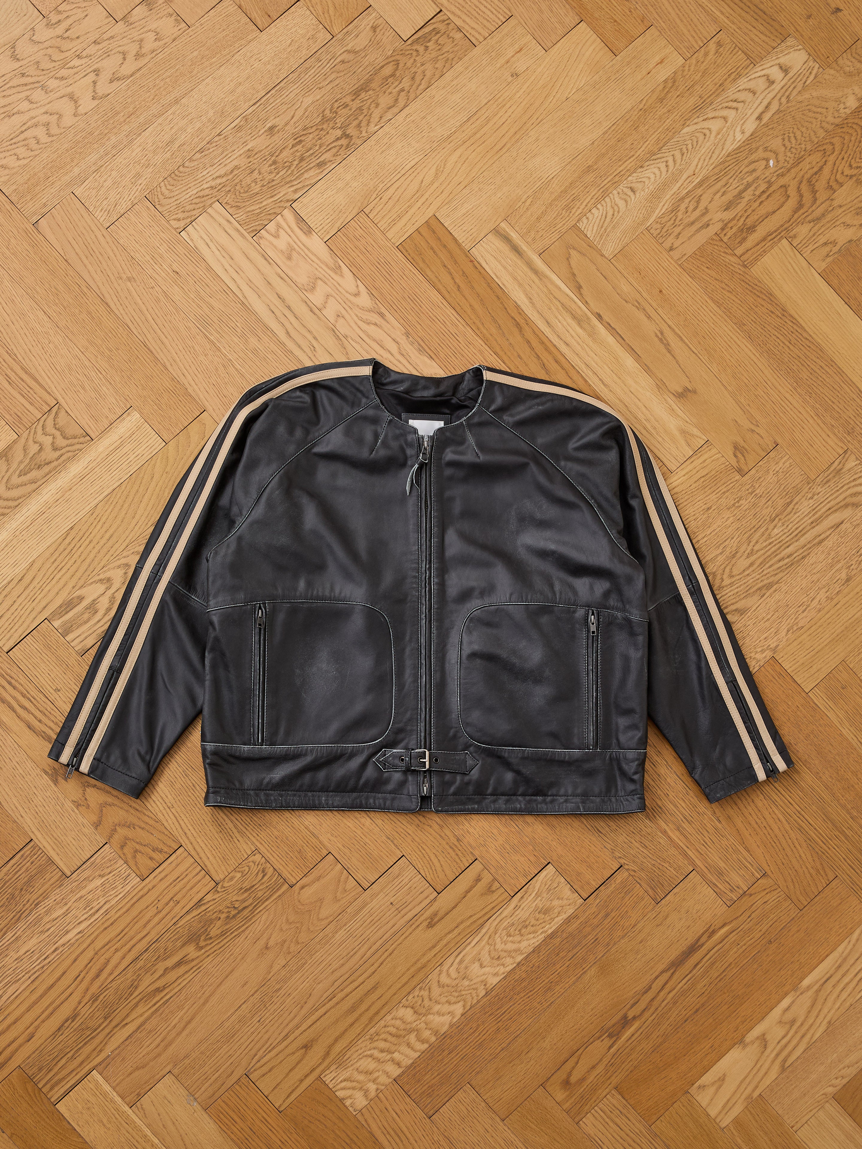 The FOUND Leather Track Jacket features black leather with beige sleeve stripes, zip front, two zippered pockets, and waist buckle, shown laid flat on a wooden herringbone floor.