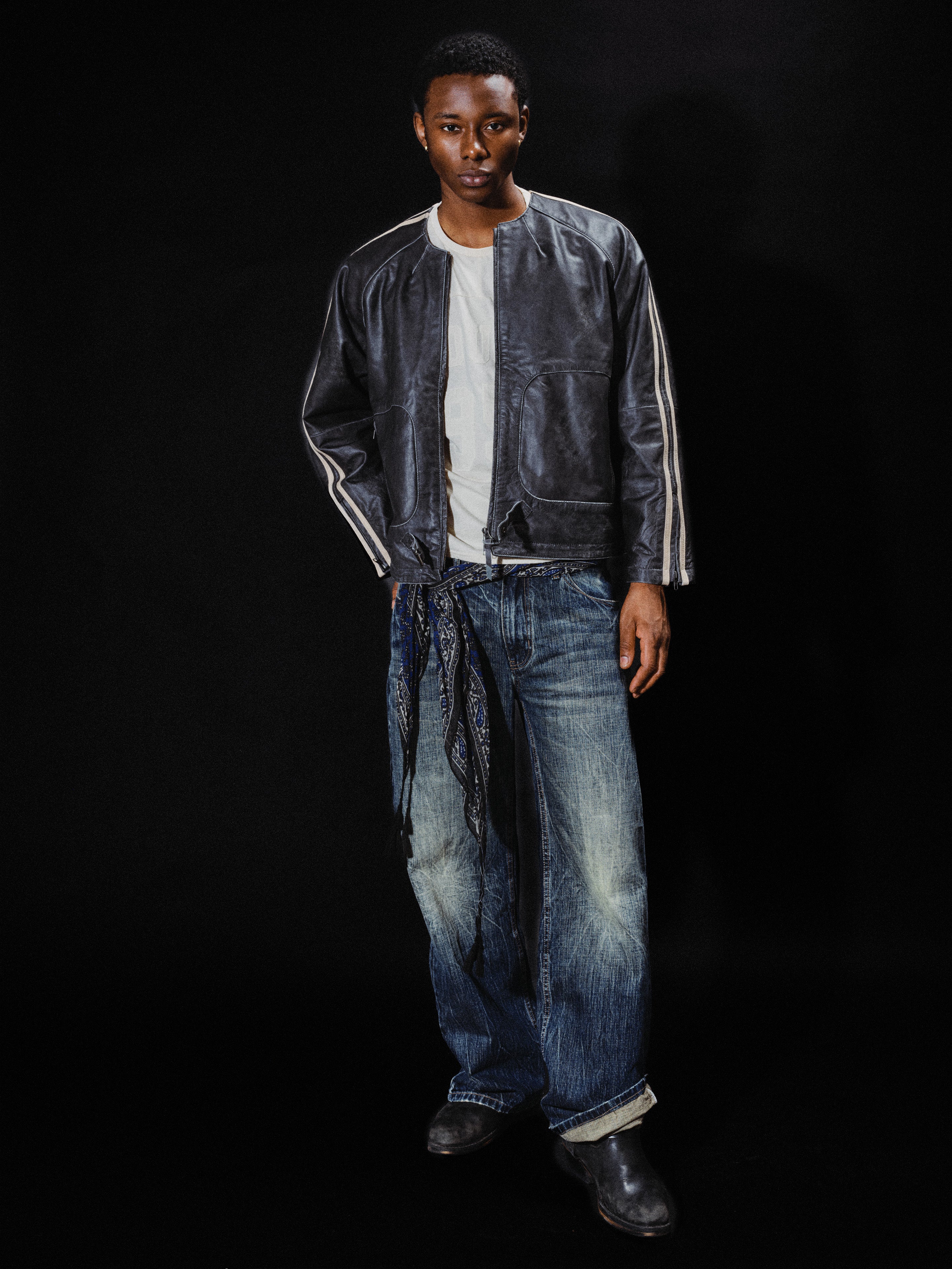 A person stands against a black background wearing the FOUND Leather Track Jacket over a white shirt, paired with blue jeans featuring a bandana tied at the waist and black boots.