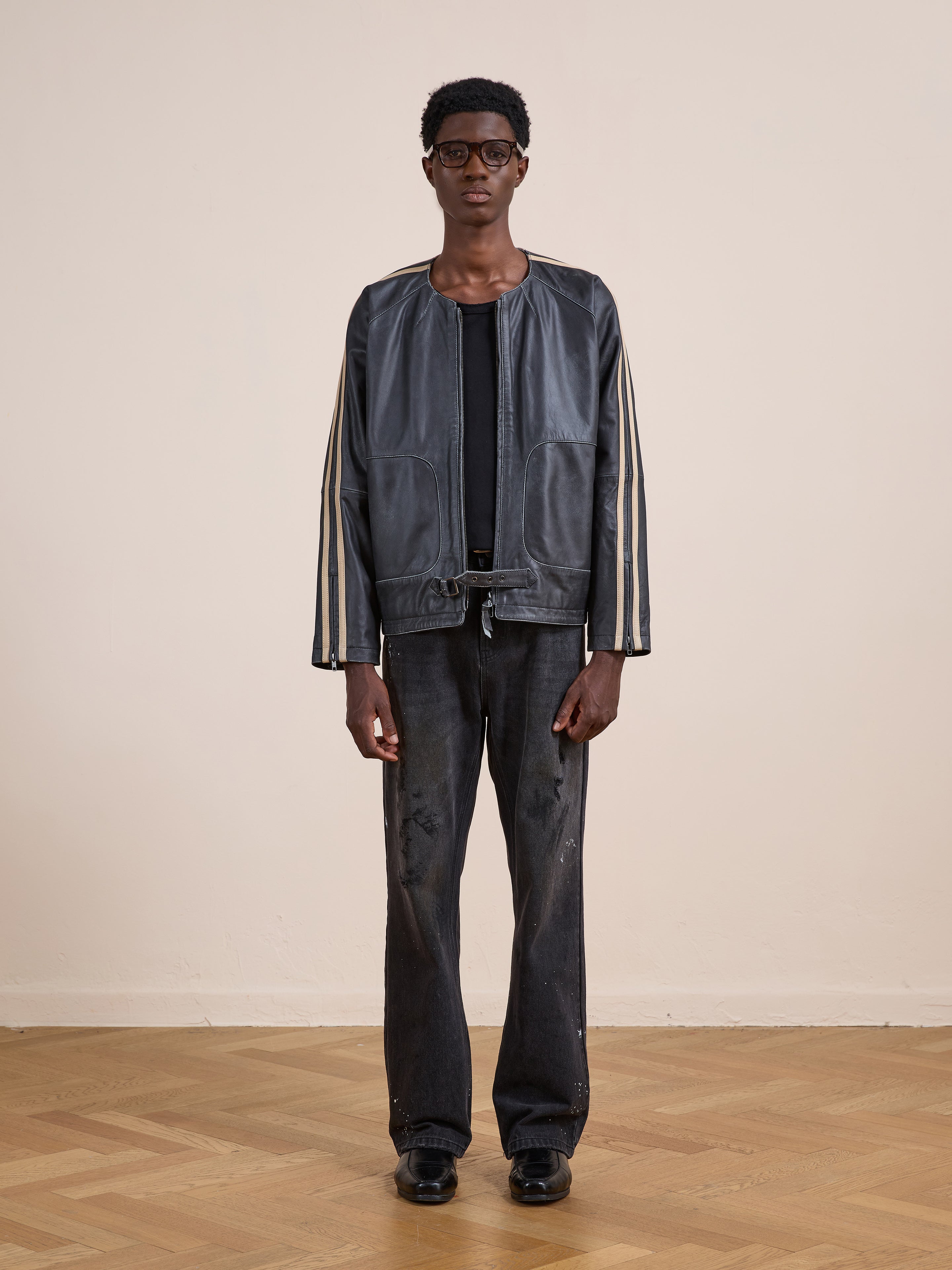 Indoors on a wooden floor, a person wears FOUND's Leather Track Jacket with white sleeve stripes, paired with a black shirt, black distressed jeans, and black shoes.
