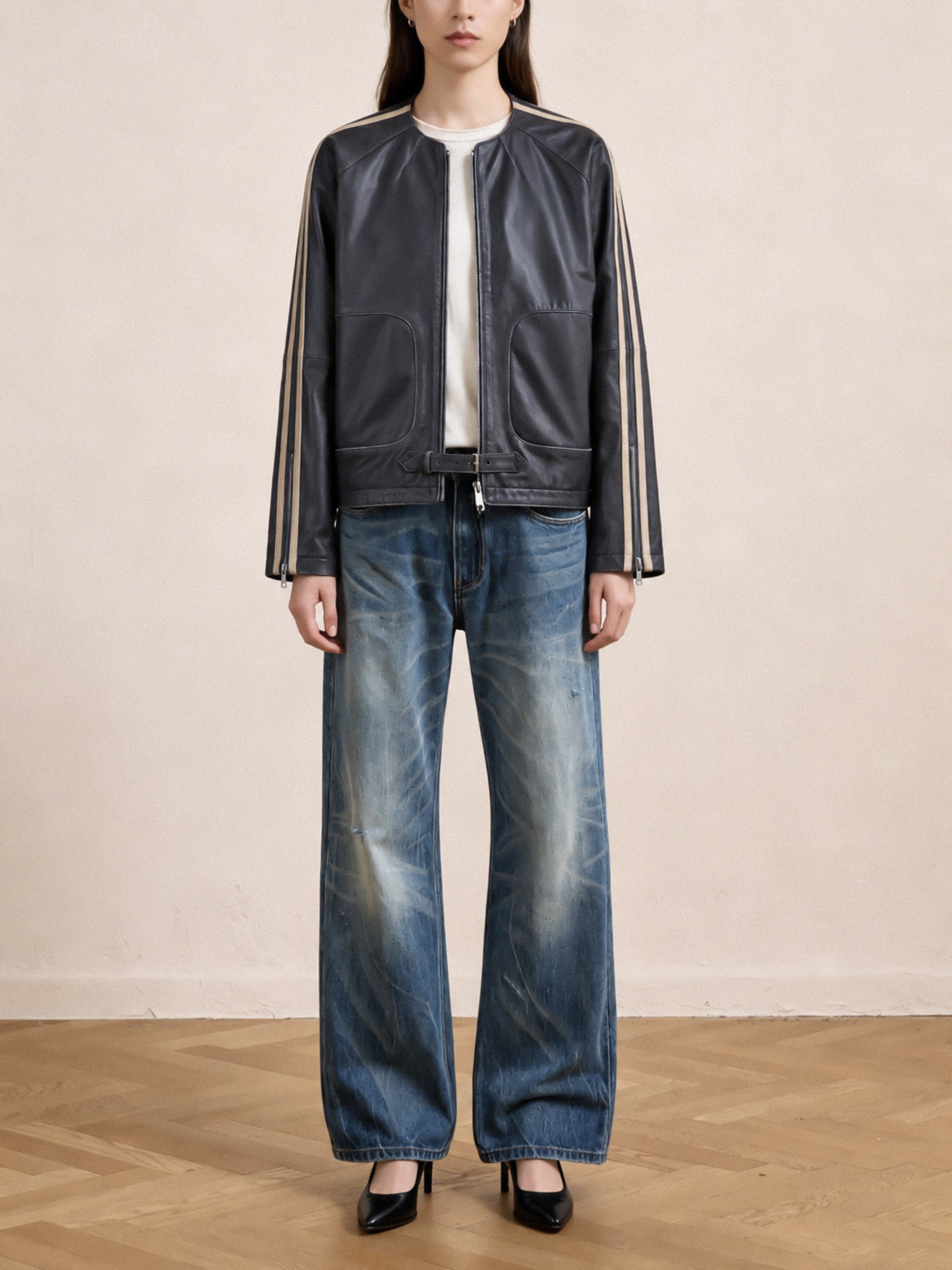 A person wearing FOUND’s Leather Track Jacket, a white shirt, wide-leg faded blue jeans, and black heels stands on a wooden floor against a plain beige wall, effortlessly channeling the timeless edge of this vintage-inspired piece.