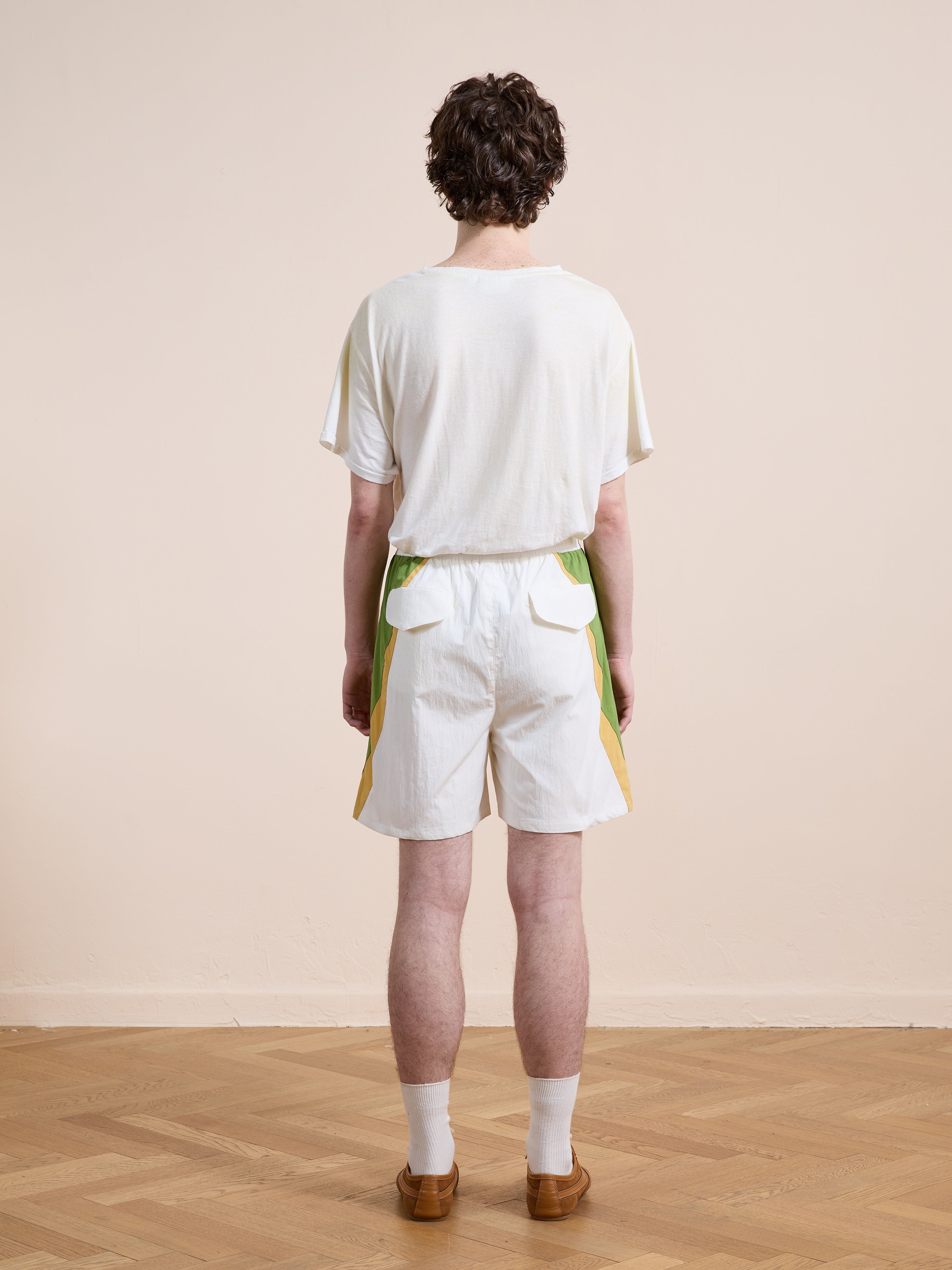 A person with short brown hair stands indoors on a wooden floor, wearing a white t-shirt and Found Legacy Cricket Track Shorts, featuring a relaxed fit with green and yellow accents, along with white socks and brown shoes.