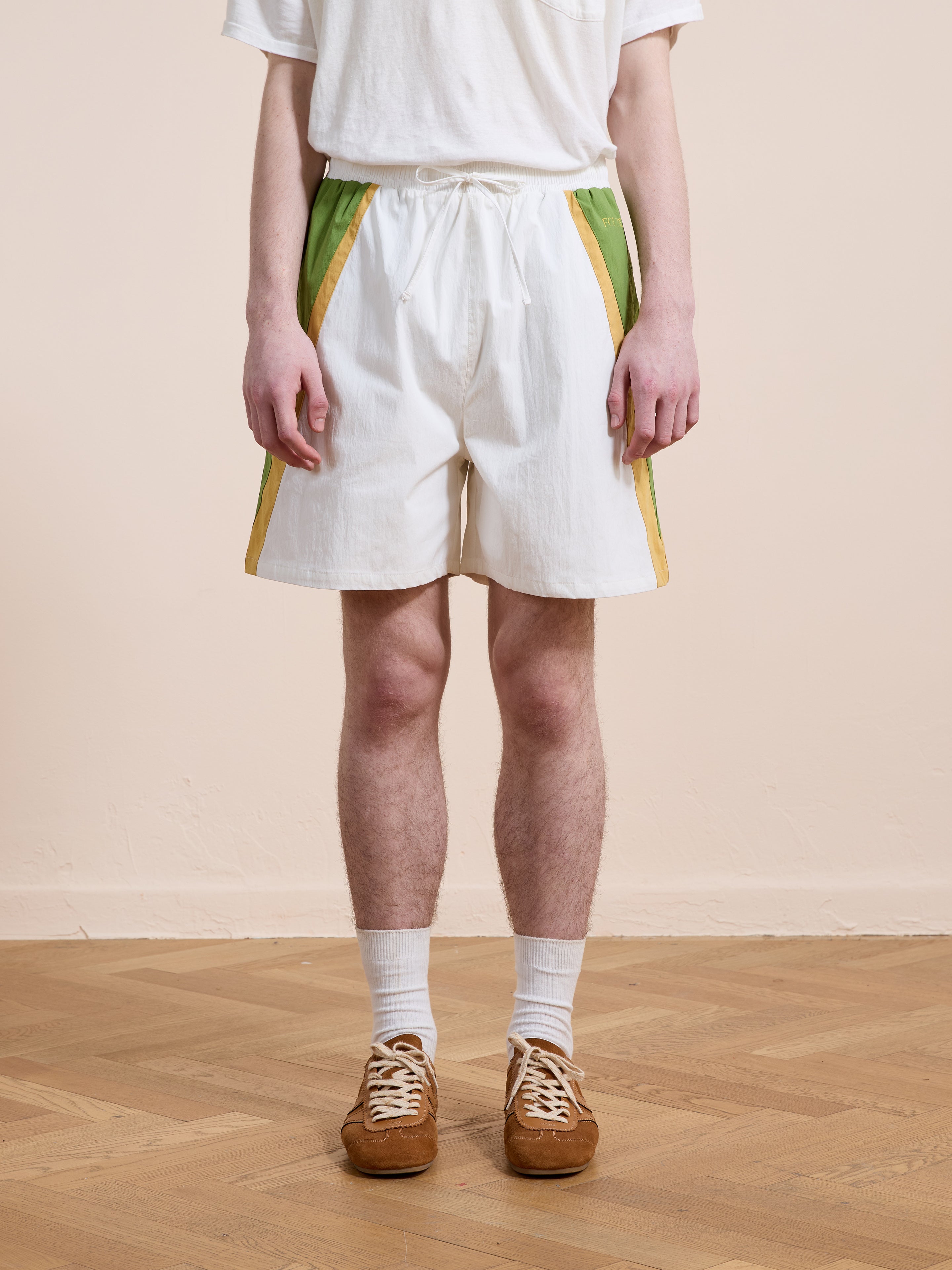A person stands indoors on a wooden floor, wearing a white t-shirt, white socks, brown sneakers, and Found Legacy Cricket Track Shorts—unisex shorts with green and yellow stripes.