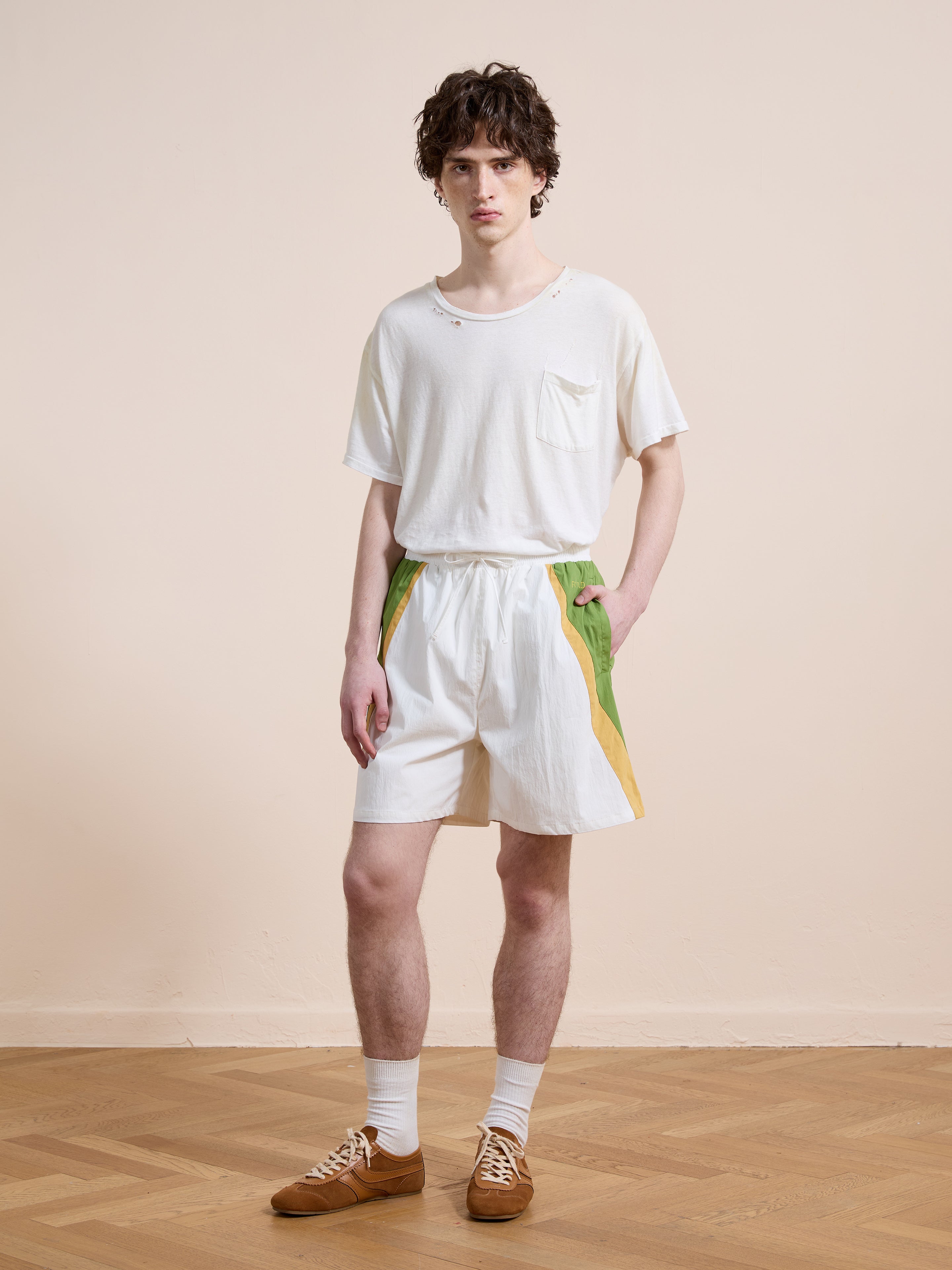 A young man stands indoors wearing a white t-shirt with small holes, Found’s Legacy Cricket Track Shorts featuring green and yellow side stripes, white socks, and brown sneakers.