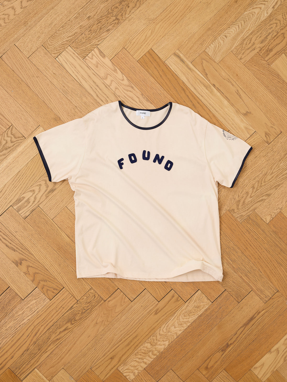 The FOUND Logo Felt Patch Ringer Tee in beige features short sleeves and navy trim on the collar and sleeves, with a navy “FOUND” felt patch logo on the front. Shown laid flat on a wooden herringbone floor, this unisex tee offers a relaxed fit for all-day comfort.