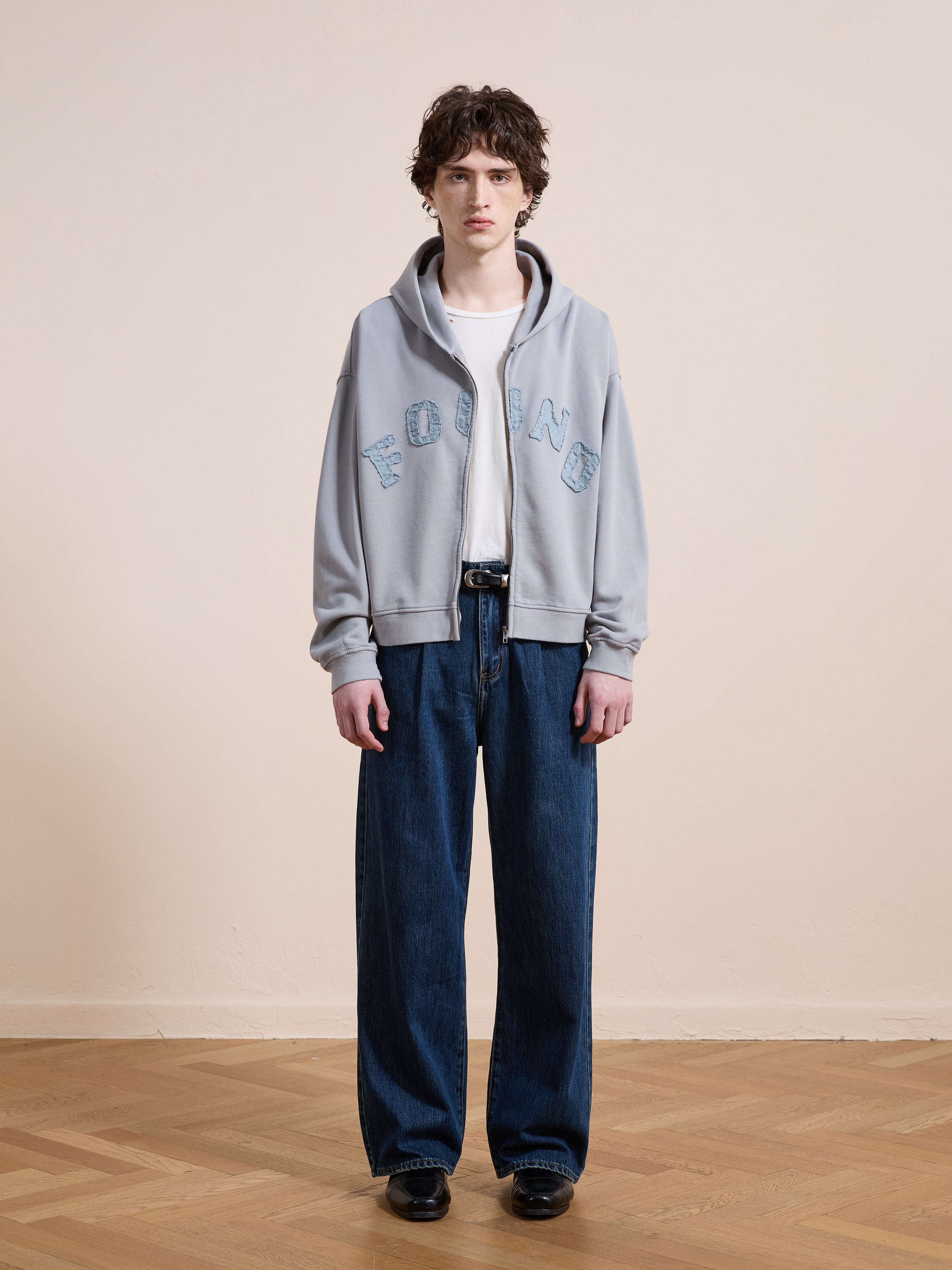 A person stands on a wooden floor against a beige wall, wearing the FOUND Logo Patch Hoodie in light gray, a white tee, unisex relaxed fit wide-leg jeans, and black shoes.