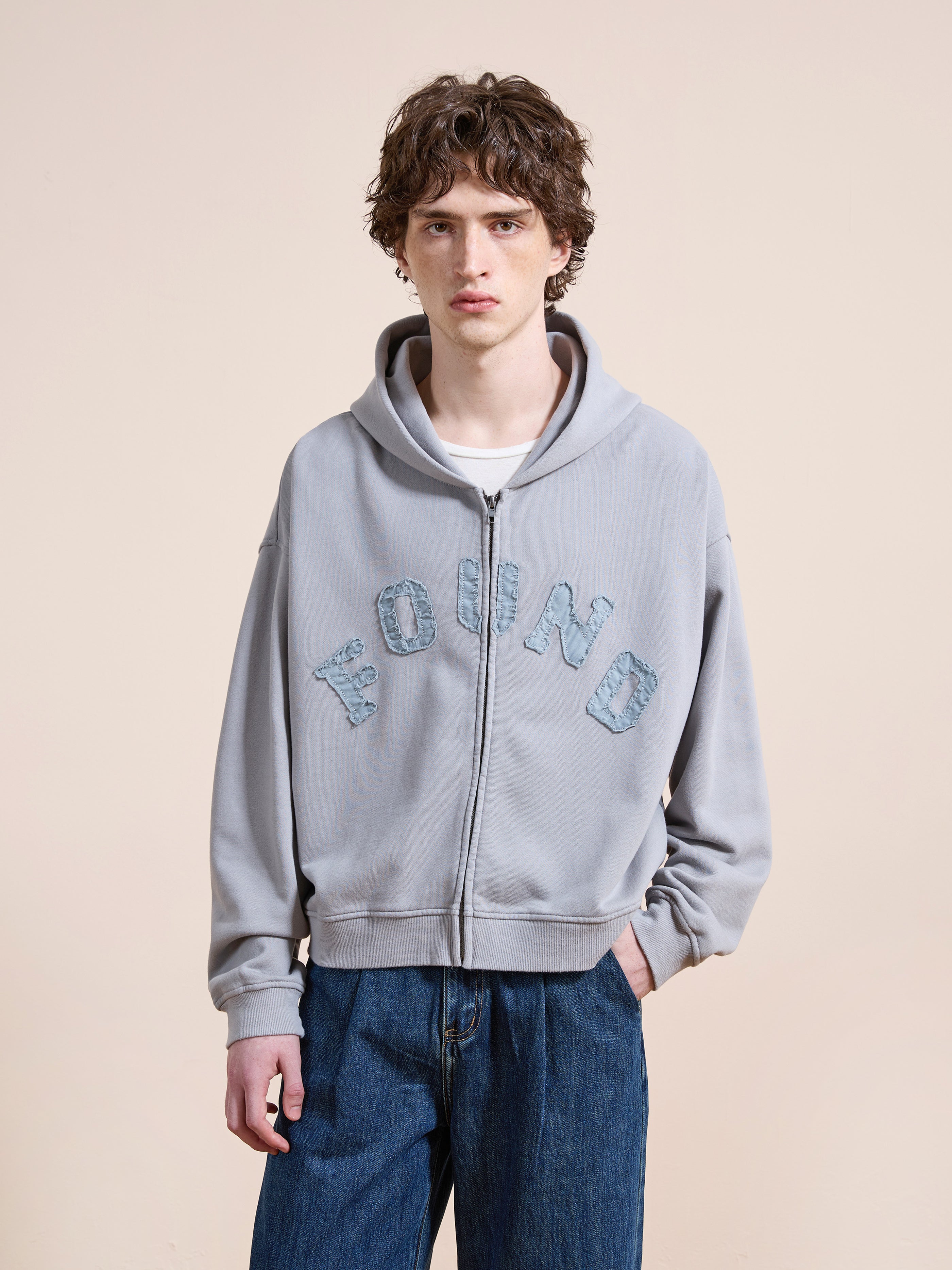 Someone wearing the FOUND Logo Patch Hoodie in light gray, a unisex relaxed-fit zip-up with textured "FOUND" lettering, is seen paired with blue jeans against a plain beige background.