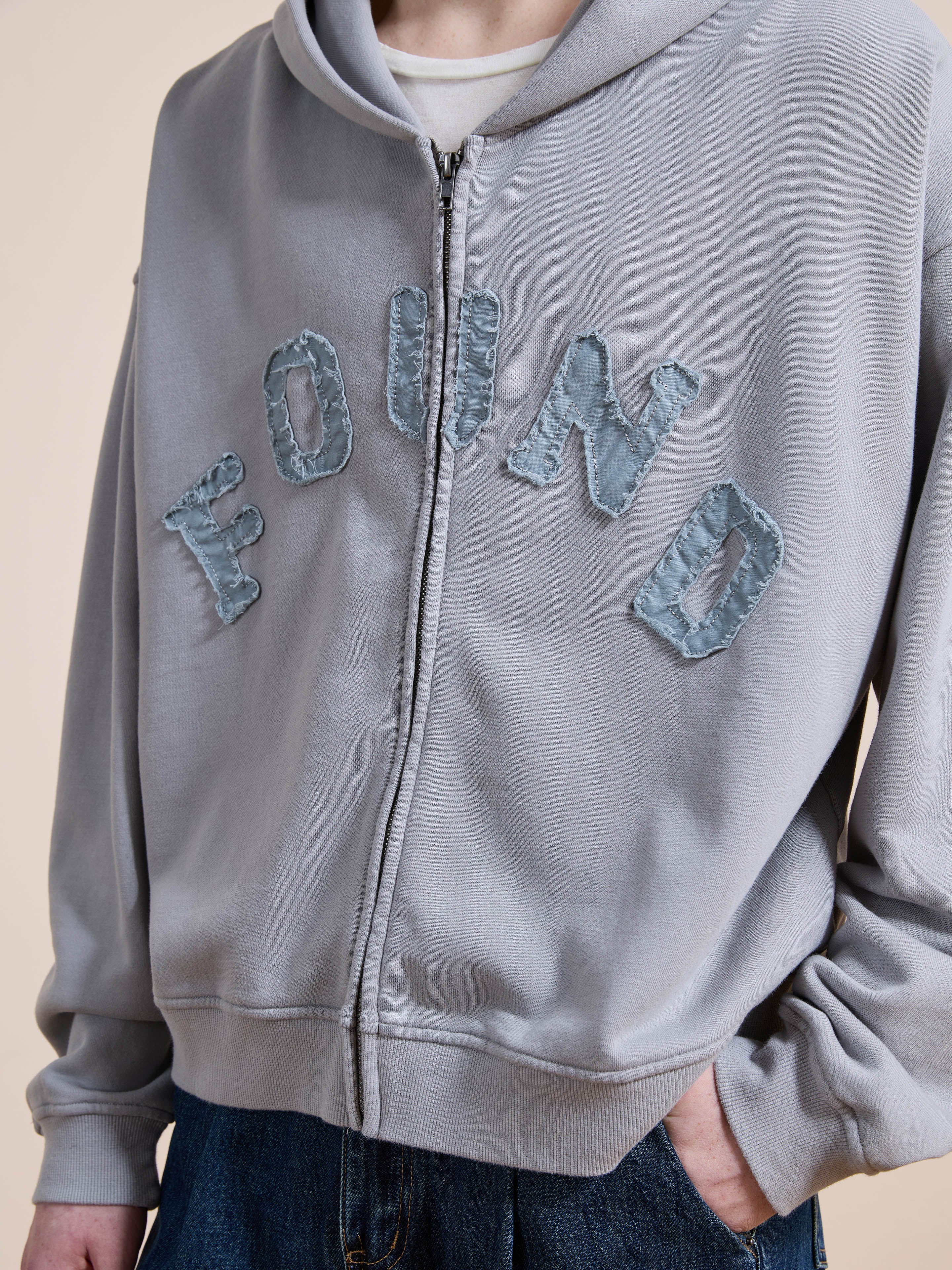 A person wearing the FOUND Logo Patch Hoodie, a light gray unisex relaxed-fit zip-up with "FOUND" stitched on the front, styled with dark jeans for a vintage athletic vibe.