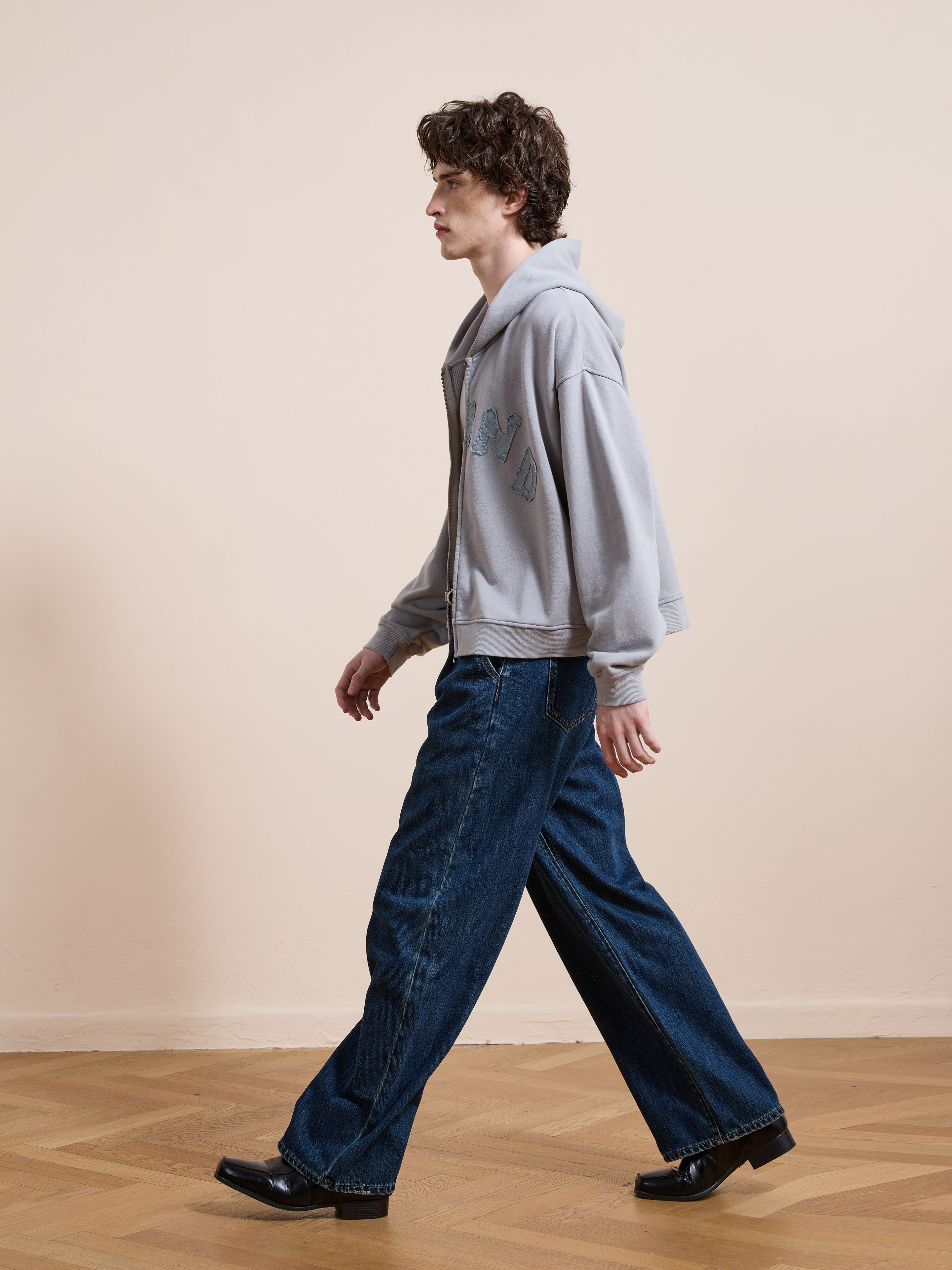 A person wearing the FOUND Logo Patch Hoodie in gray and wide-leg unisex jeans walks indoors on a wooden floor against a plain beige wall.