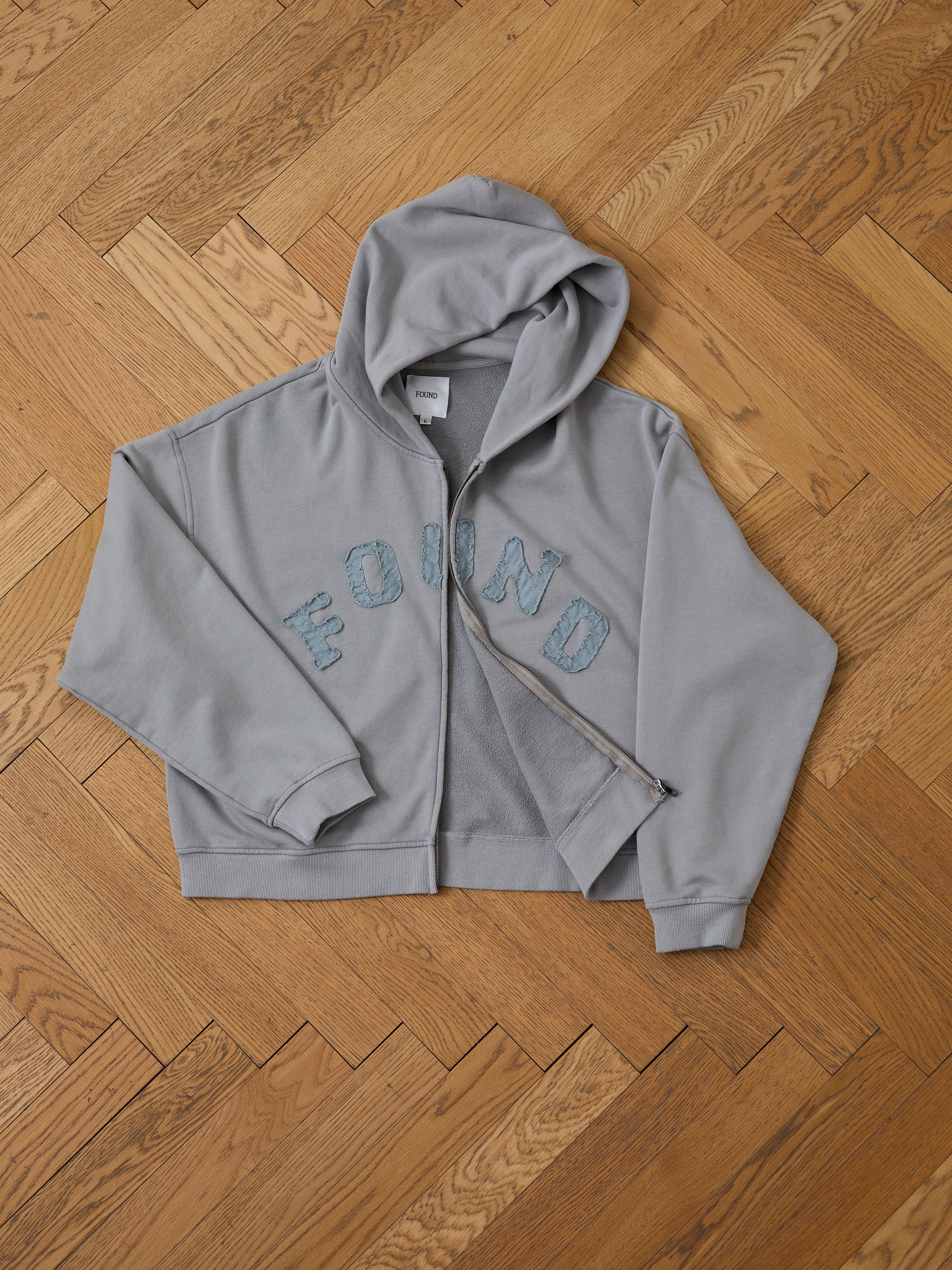 The FOUND Logo Patch Hoodie, a gray unisex zip-up with blue textured “FOUND” lettering on the front, is displayed laid flat on a wooden herringbone floor for a vintage athletic vibe.