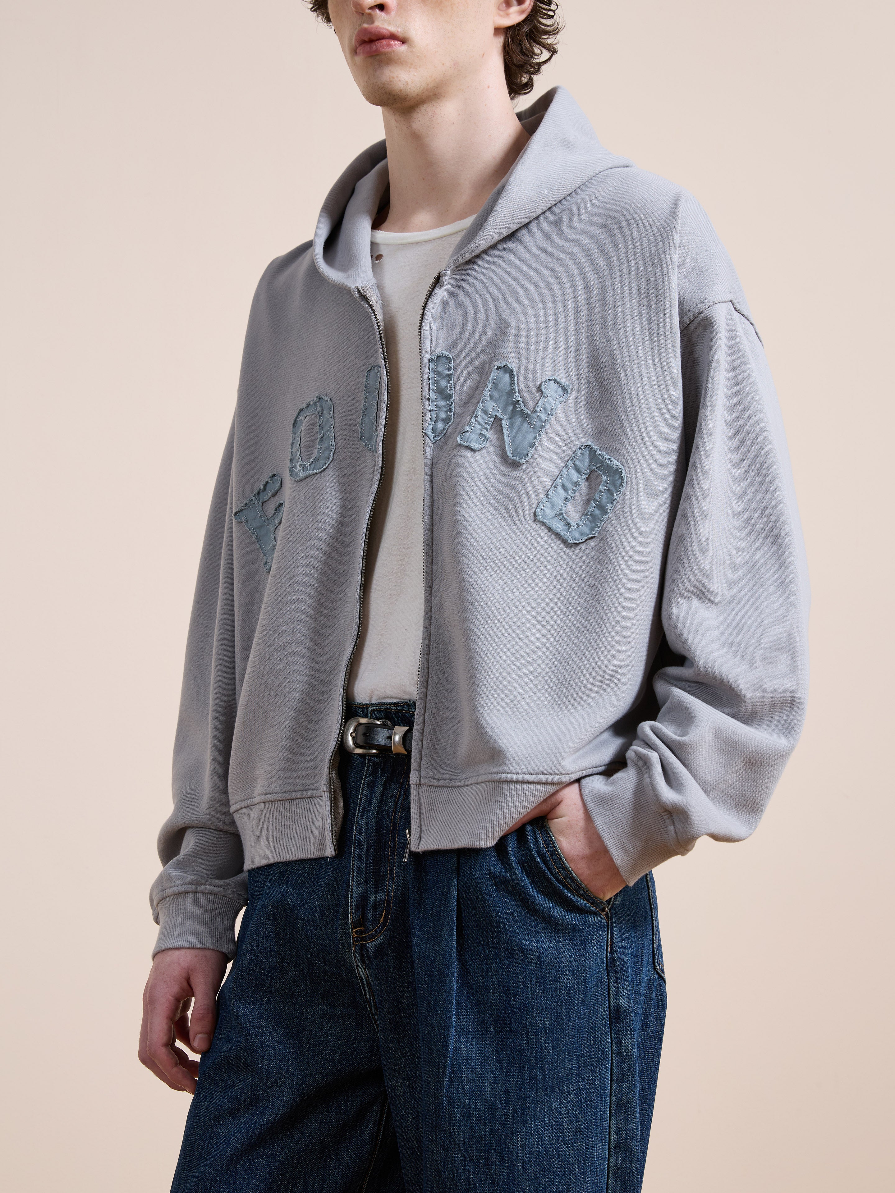 A person wears the FOUND Logo Patch Hoodie—a washed gray, unisex relaxed-fit zip-up—layered over a white t-shirt and blue jeans for a vintage athletic style.