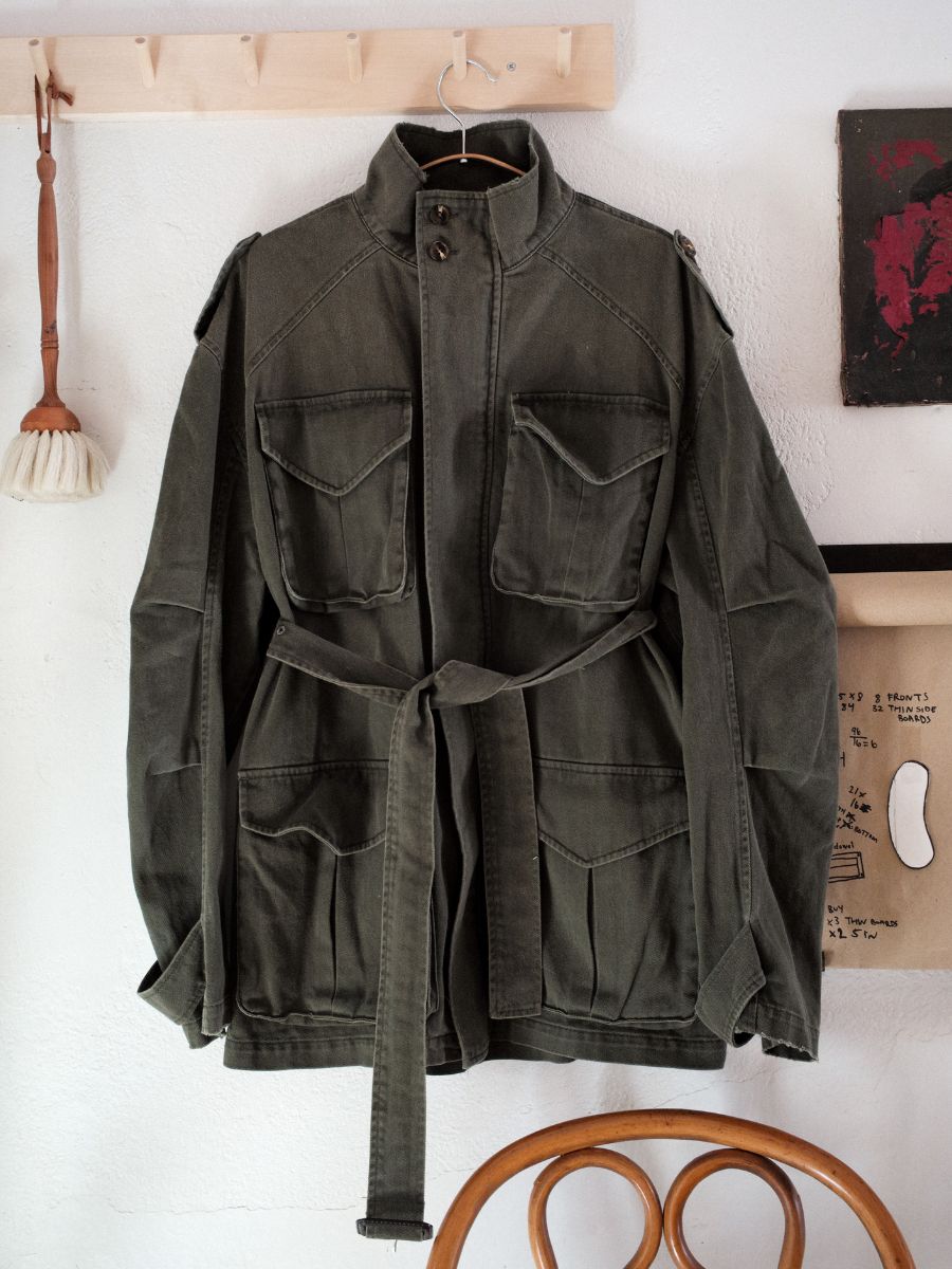 The M65 Utility Twill Pocket Jacket by FOUND, with its olive green color and belt, hangs on a wooden peg against a white wall. Its military-inspired design with multiple pockets is artistically complemented by a nearby brush and abstract art piece.