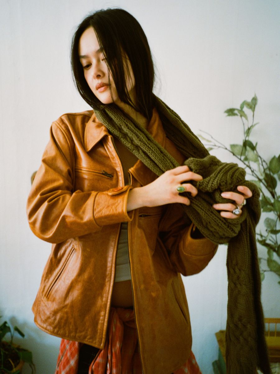 A woman in a brown leather jacket adjusts her FOUND Forest Distressed Cable Knit Scarf. With long dark hair and multiple rings, she stands with a plant and small furniture piece in the background.