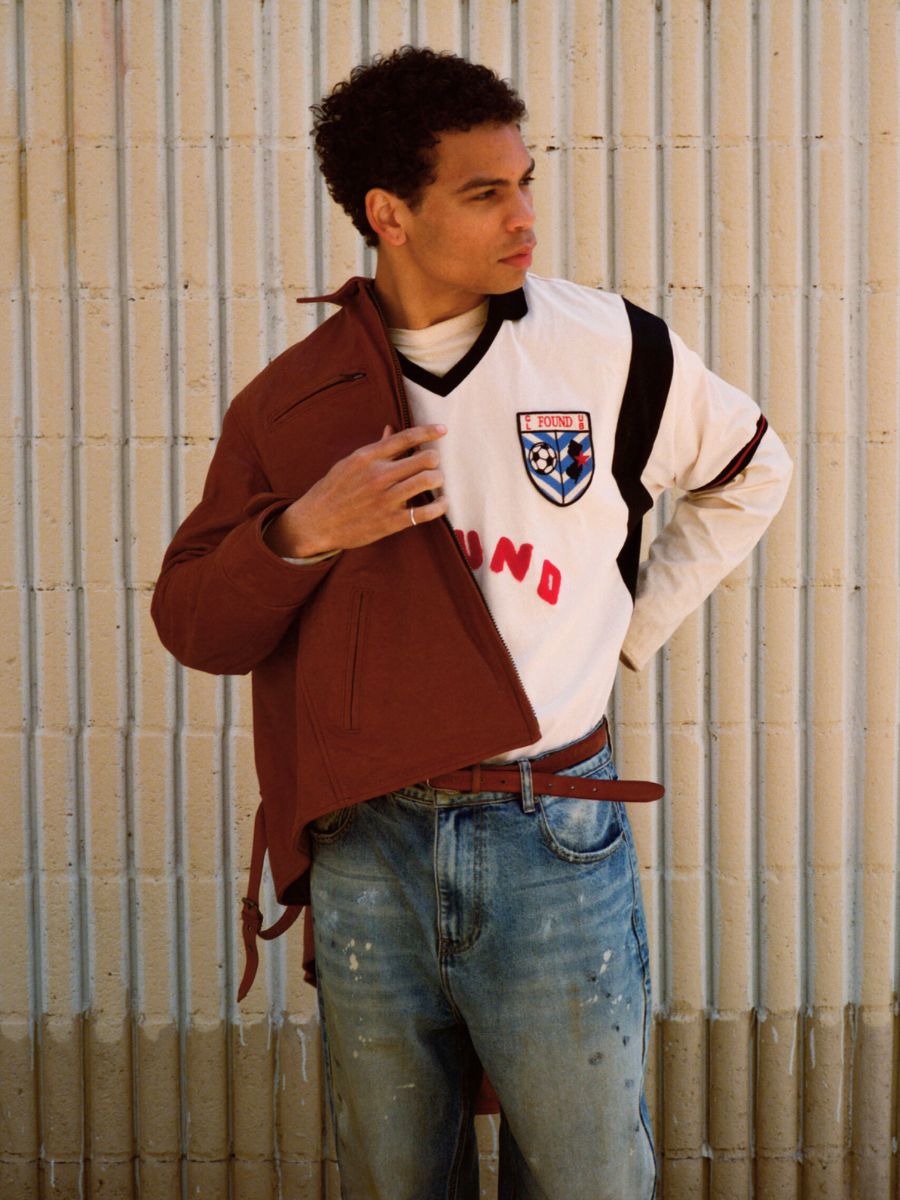 Wearing a FOUND Marlboro Sun Faded Leather Racer Jacket over a white soccer jersey and blue jeans, a person stands against a textured beige wall, gazing to the side.