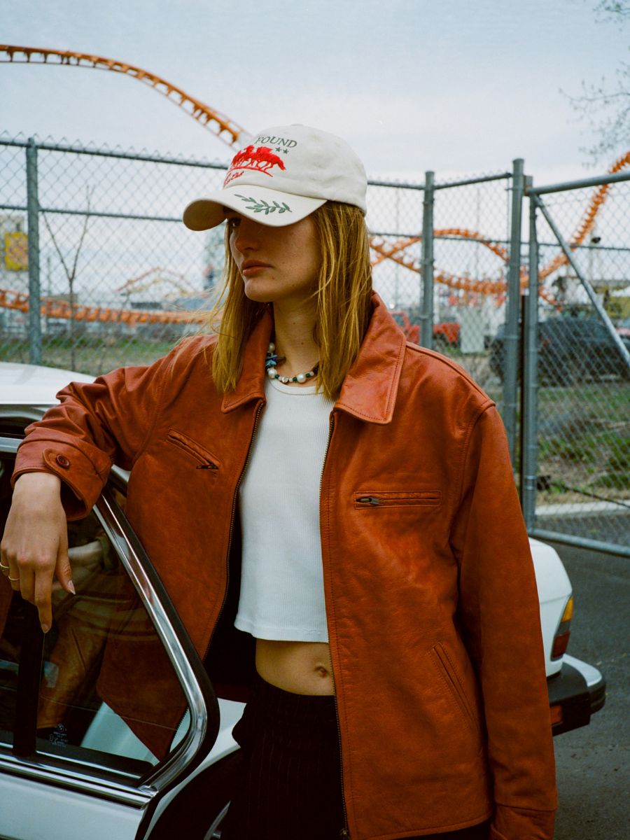 A person wearing a tan baseball cap, white crop top, and FOUND Marlboro Sun Faded Leather Racer Jacket stands by a car with an amusement park roller coaster in the background.