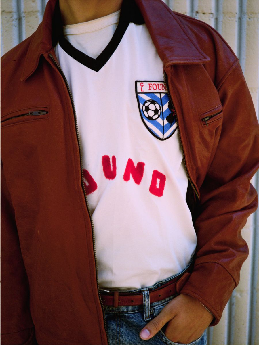 A person wearing the FOUND Marlboro Sun Faded Leather Racer Jacket over a white soccer jersey with a crest and red letters, paired with blue jeans and a brown belt.