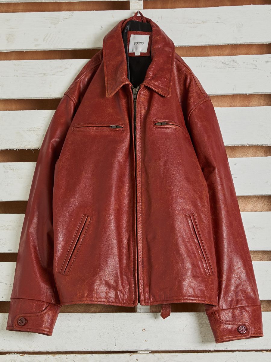A red Marlboro Sun Faded Leather Racer Jacket by FOUND, perfect as a unisex leather trucker, features two front zippers, two lower pockets, and a collared design. It's displayed on a white wooden slatted wall.