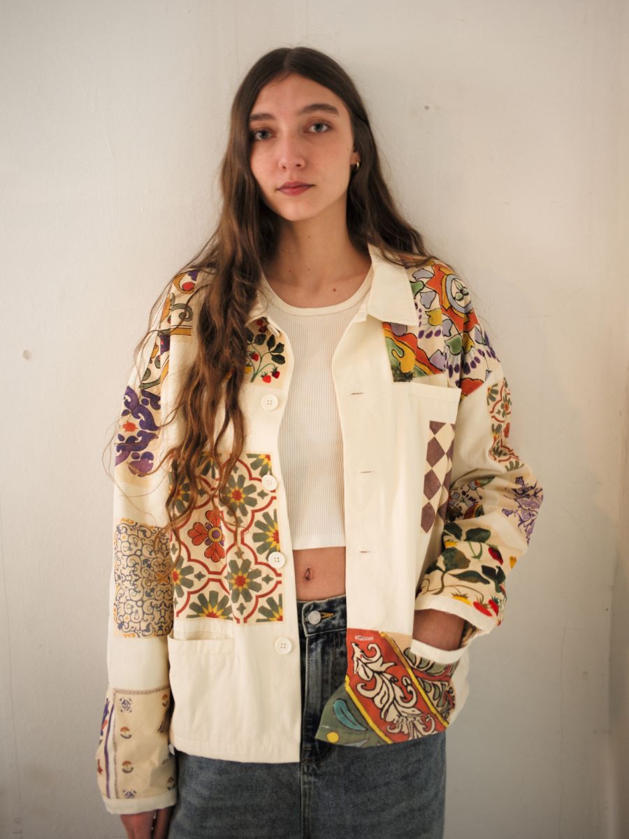 A woman with long hair wears a vibrant FOUND Mosaic Work Jacket over a white top and jeans, standing against a plain wall.