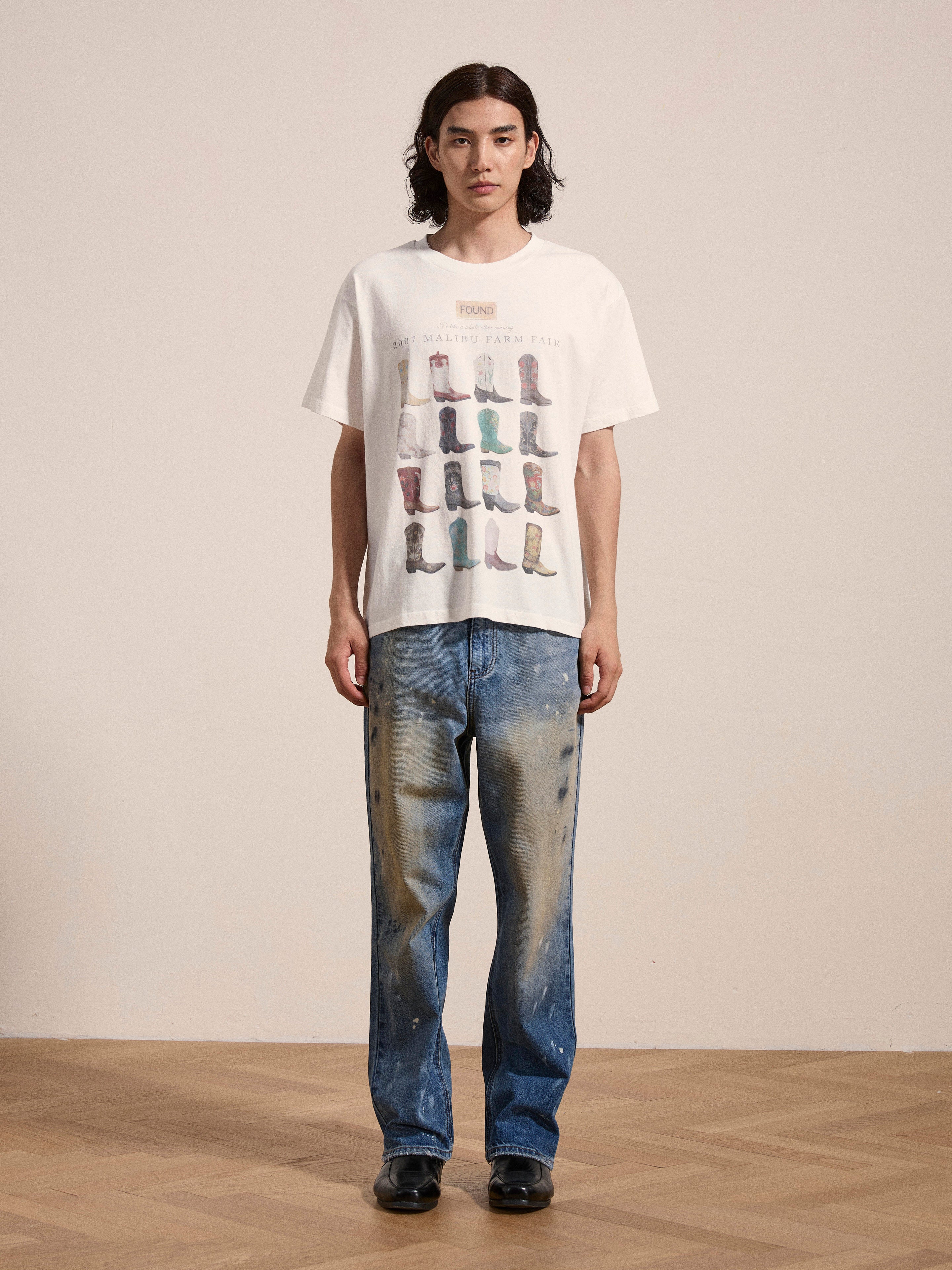 A person with long hair is wearing a 100% cotton Malibu Farm Fair Tee by FOUND, which showcases various boots. They are paired with distressed blue jeans and black shoes, standing on a wooden floor against a plain wall.