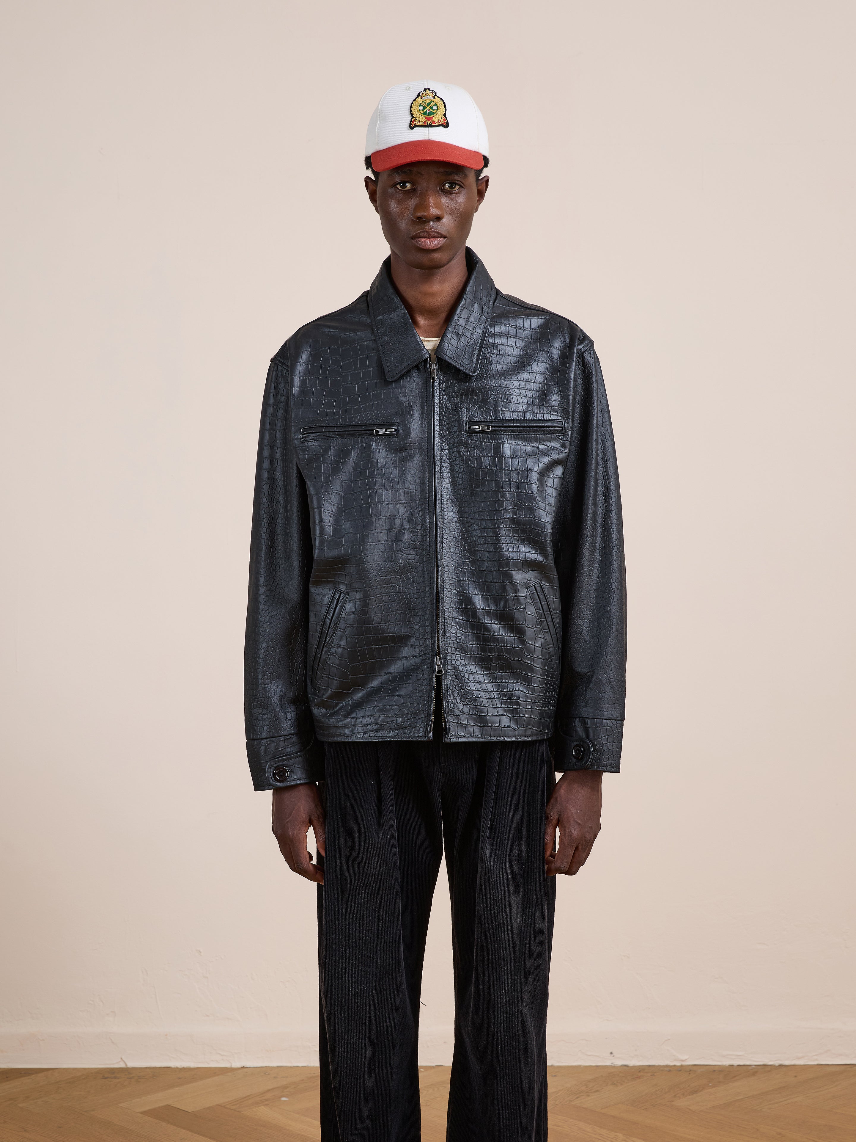 A person wearing the FOUND Marlboro Croc Embossed Leather Racer Jacket, black pants, and a white cap with a cartoon logo faces the camera against a plain beige background.