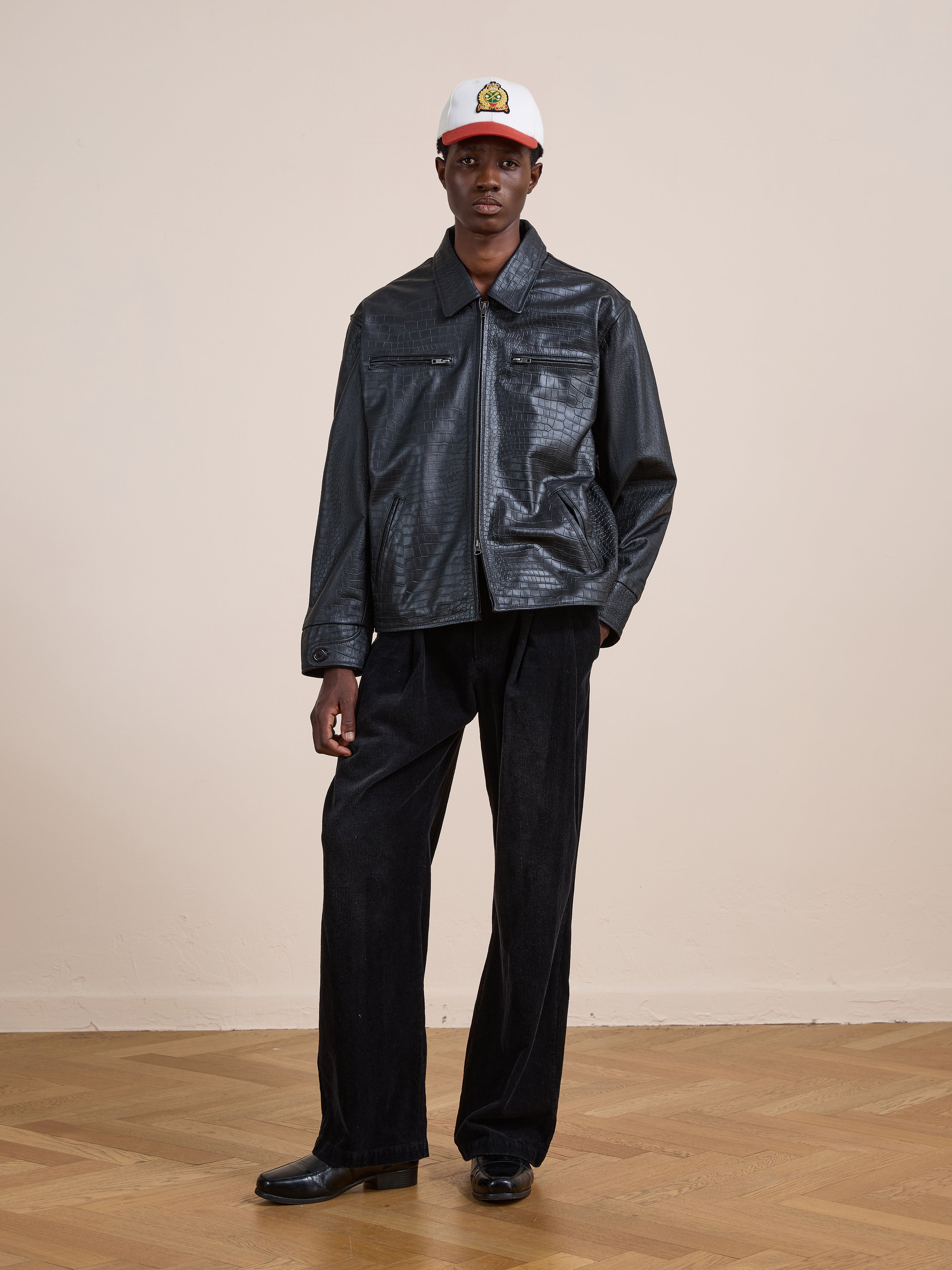 Person standing indoors on a wooden floor, wearing the FOUND Marlboro Croc Embossed Leather Racer Jacket in a unisex relaxed fit, paired with black pants, black shoes, and a white logo cap, posed against a plain beige wall.