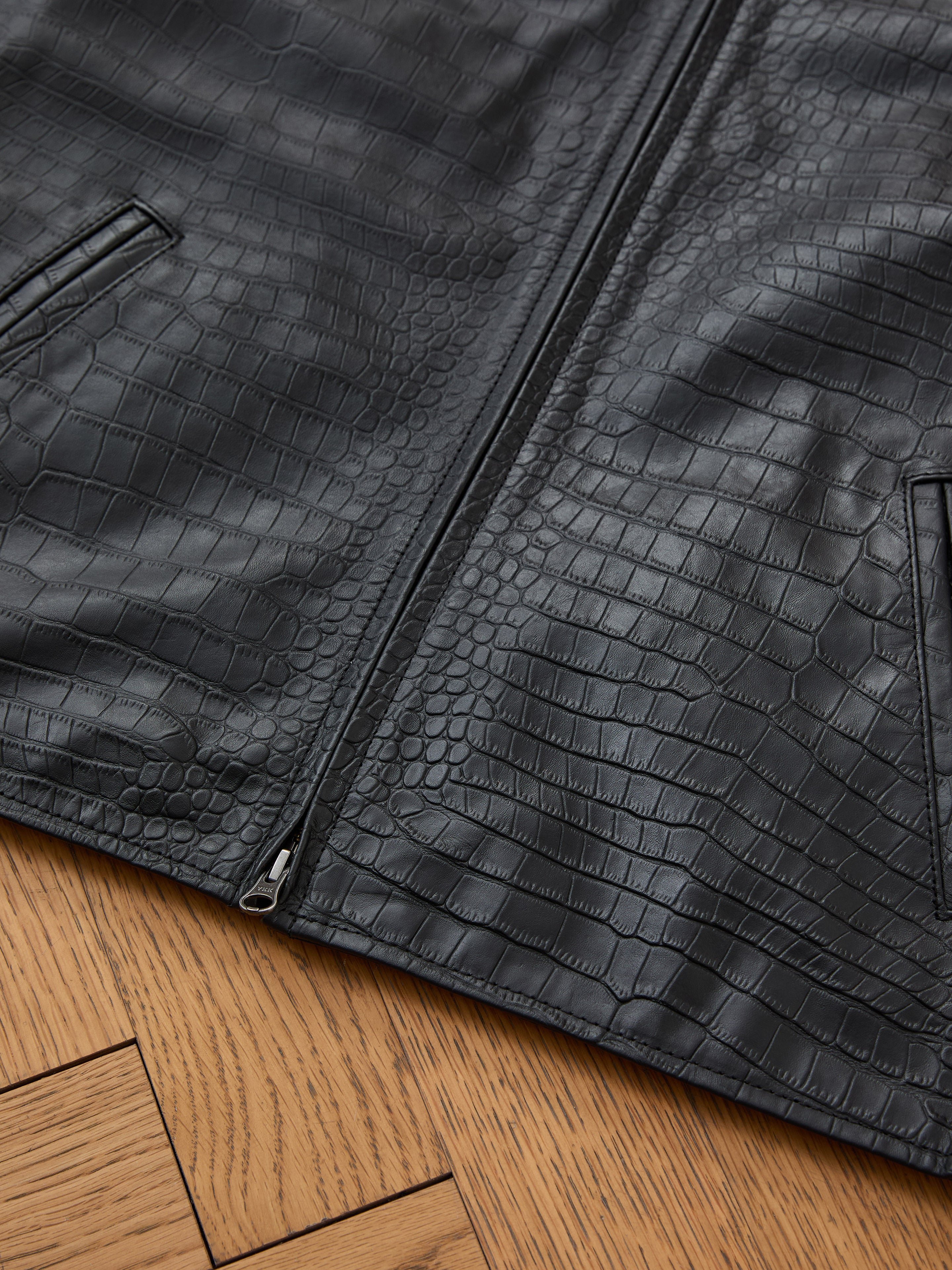 Close-up of the FOUND Marlboro Croc Embossed Leather Racer Jacket, a unisex black faux leather piece with crocodile pattern, front zipper, and two angled welt pockets, laid flat on a wooden floor.