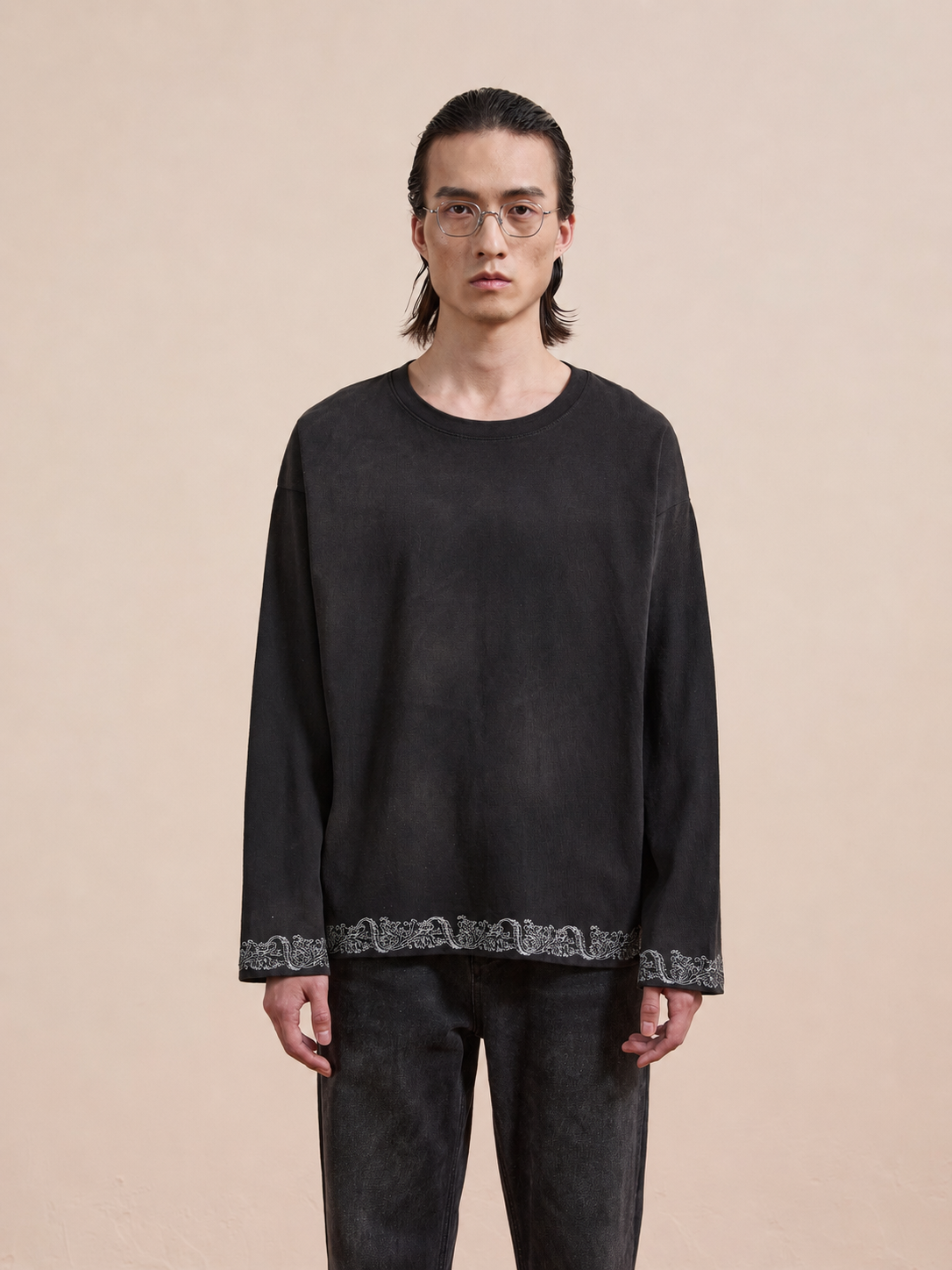A person with long dark hair and glasses wears the FOUND Metallic Long Sleeve Embroidered Tee, a unisex relaxed fit top, standing against a plain beige background.