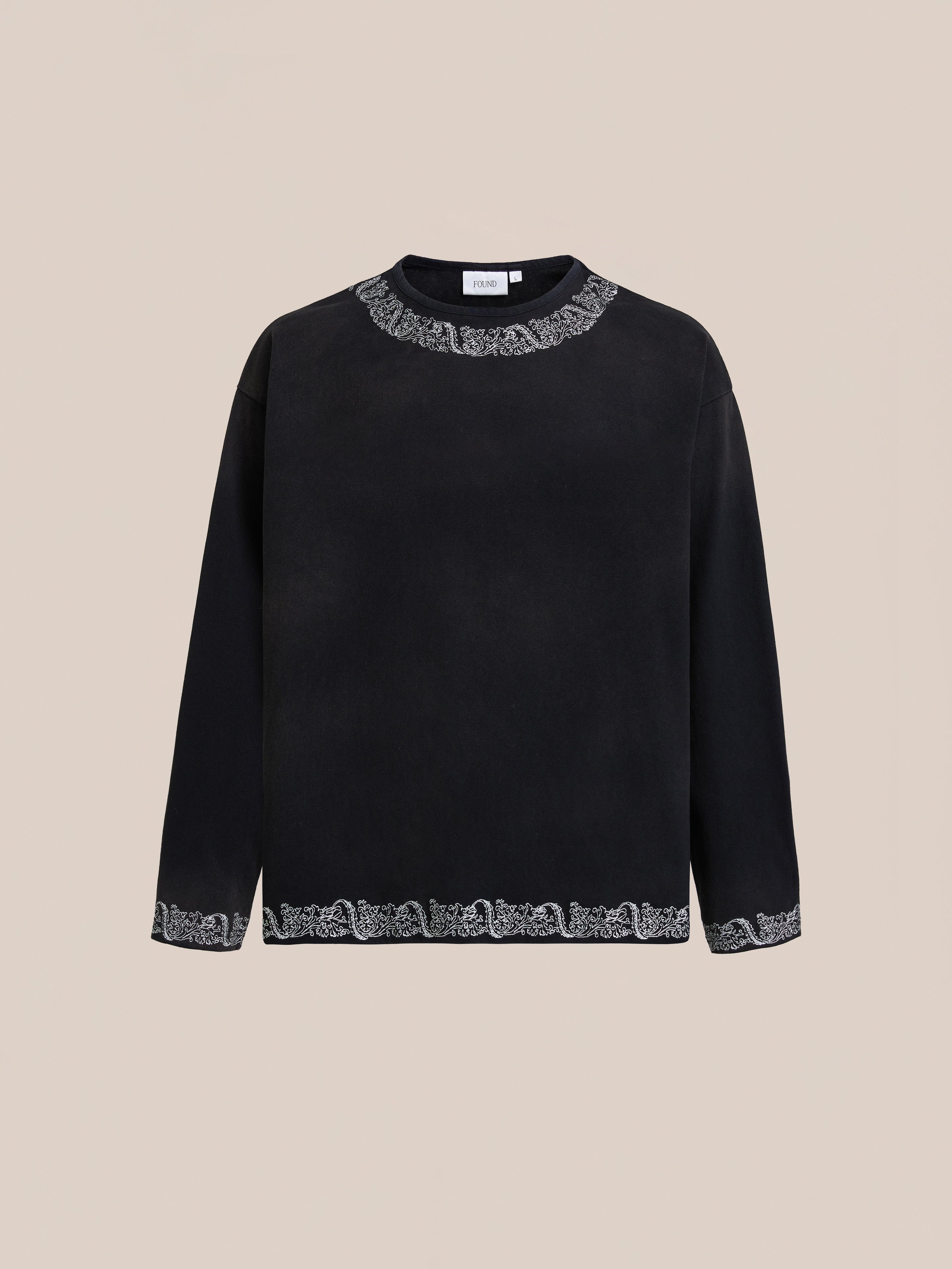 The FOUND Metallic Long Sleeve Embroidered Tee features metallic embroidery on the collar, cuffs, and hem. This unisex, relaxed-fit vintage black top is showcased against a plain beige background.