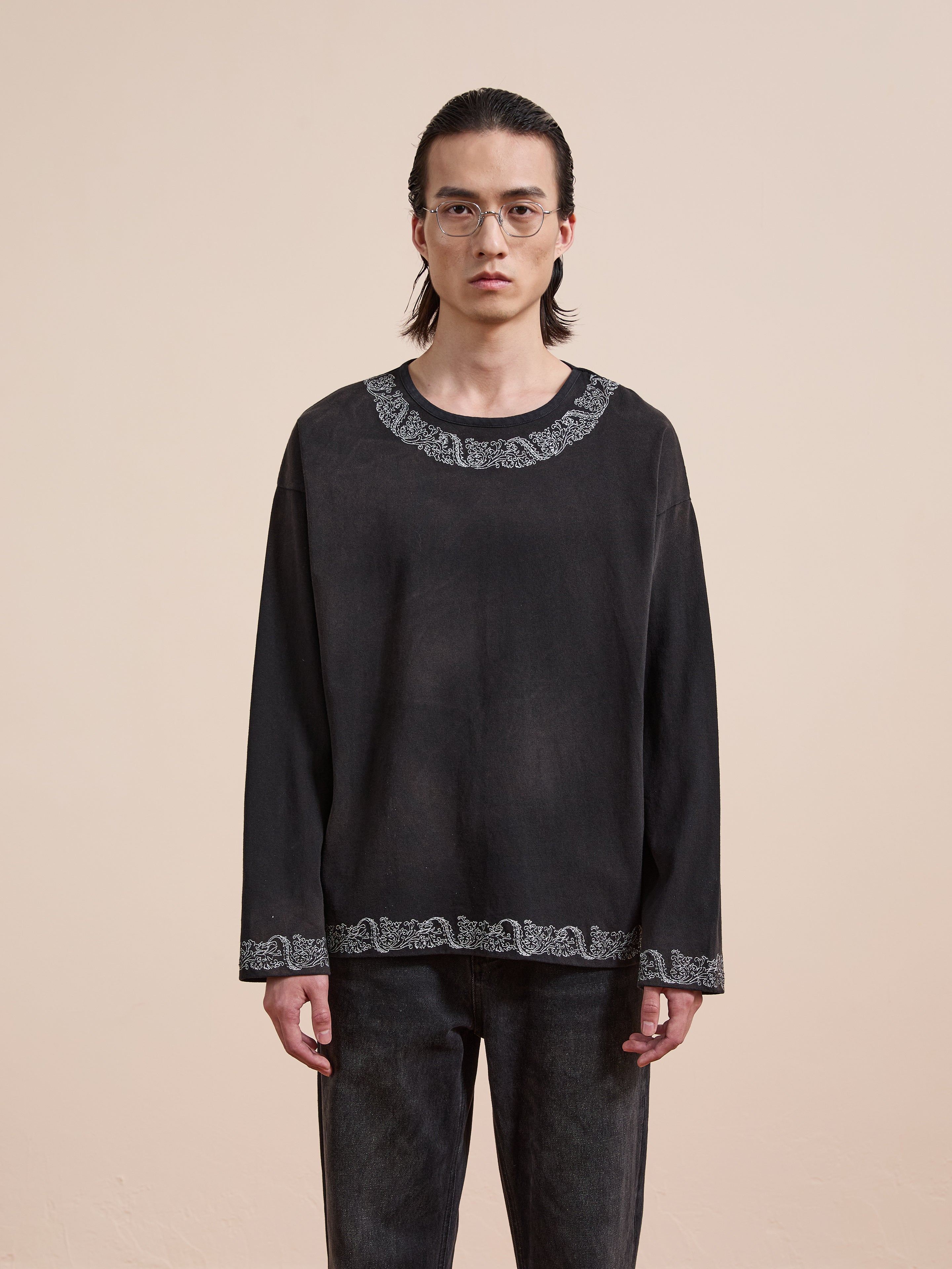 A person with long dark hair and glasses wears the FOUND Metallic Long Sleeve Embroidered Tee, featuring white embroidery on the collar and hem, against a plain beige background.