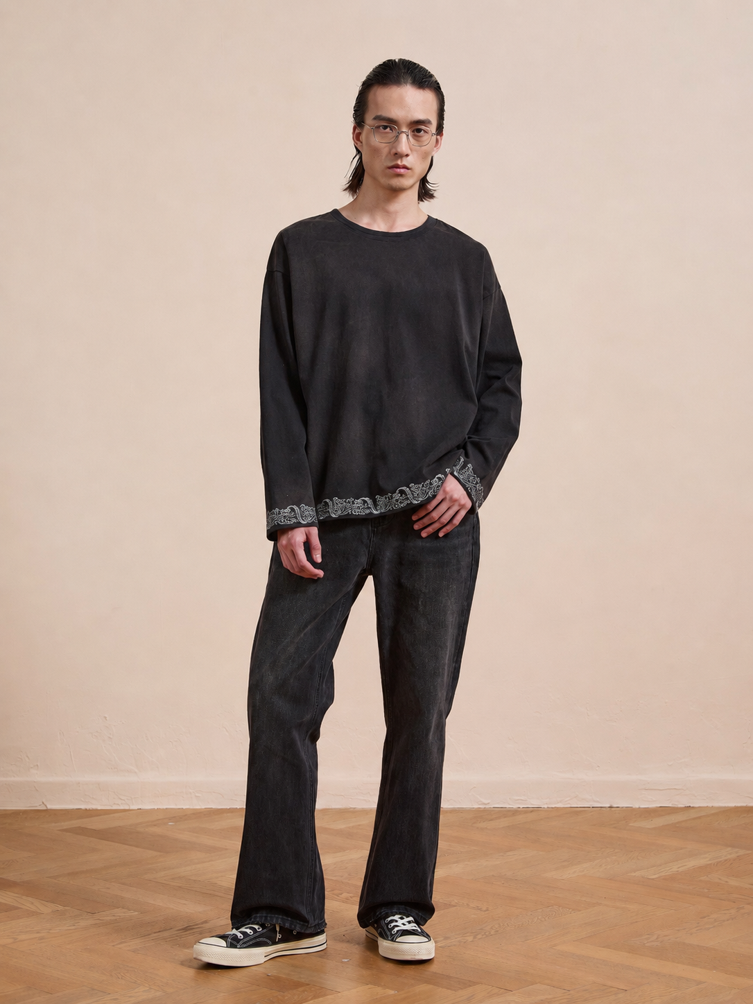 A person stands indoors on a wooden floor, facing the camera in a neutral expression while wearing the FOUND Metallic Long Sleeve Embroidered Tee with dark jeans and black-and-white sneakers.