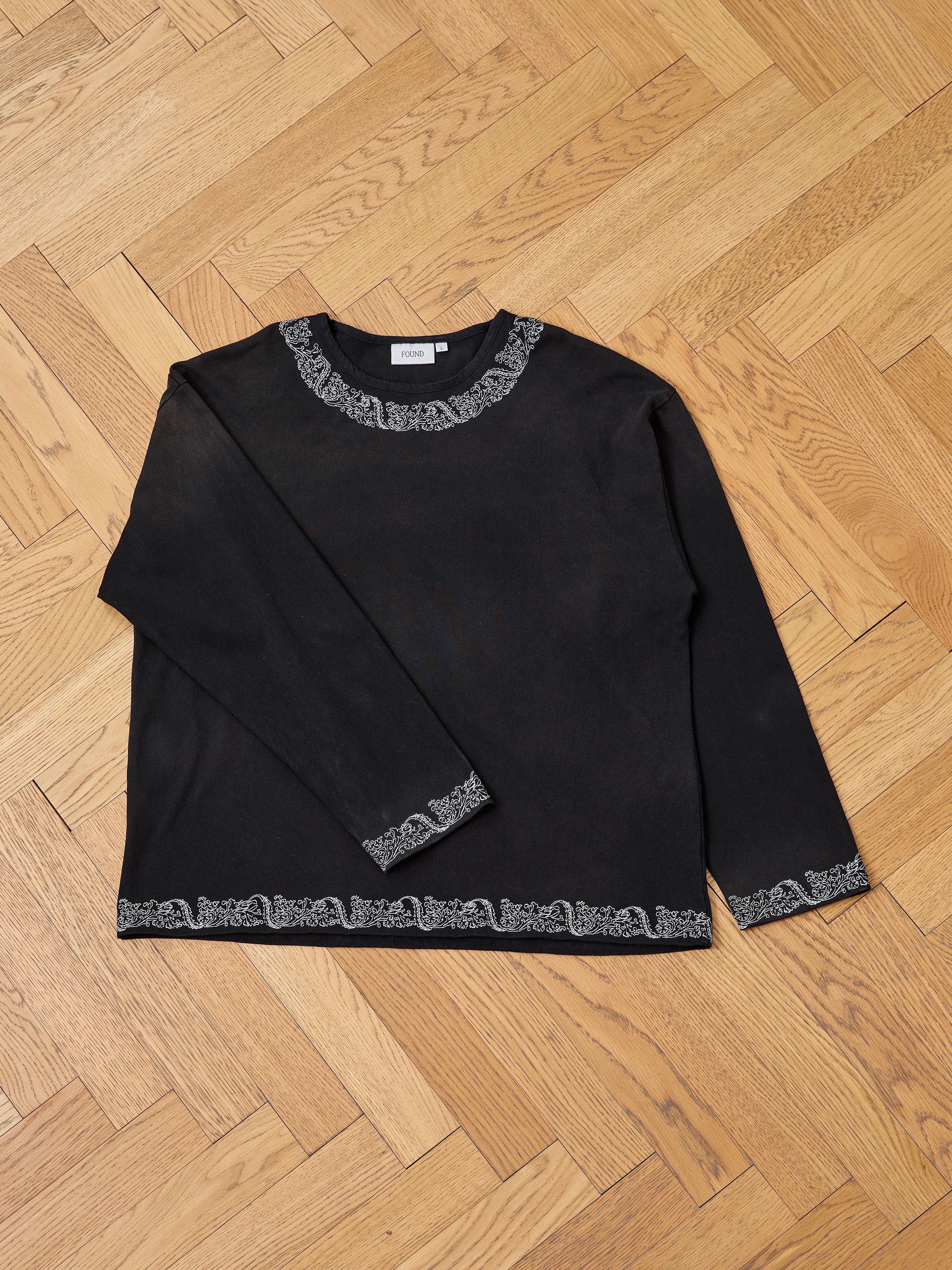 The FOUND Metallic Long Sleeve Embroidered Tee features a relaxed unisex fit in vintage black with light patterned trim on the neckline, cuffs, and hem, shown laid flat on a wooden parquet floor.