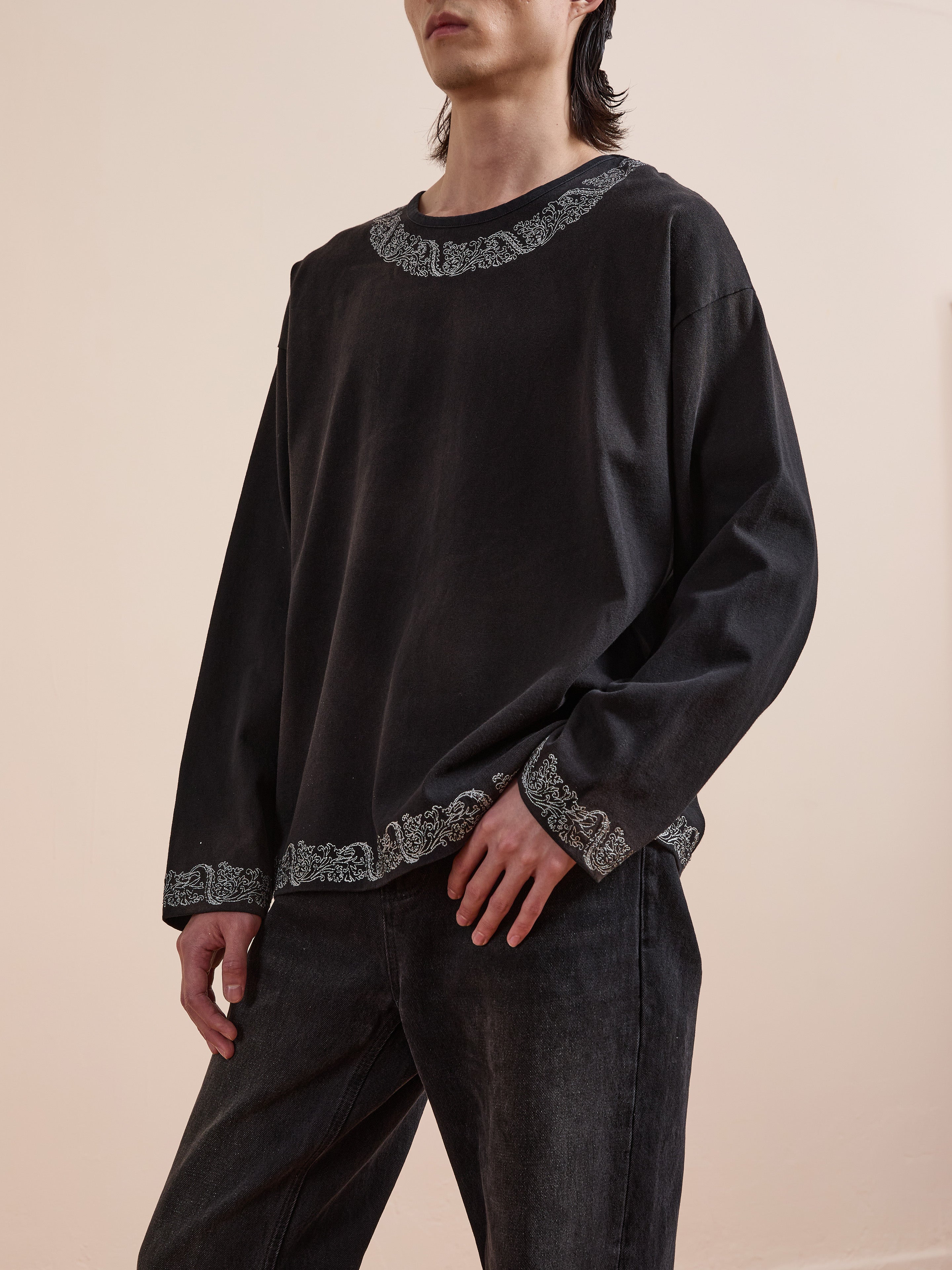 A person wearing the FOUND Metallic Long Sleeve Embroidered Tee, a vintage black unisex relaxed-fit shirt with patterned trim, paired with black jeans.