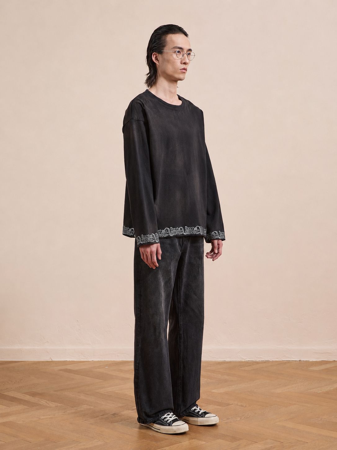 A person with long hair and glasses stands indoors wearing the FOUND Metallic Long Sleeve Embroidered Tee in a unisex relaxed fit, paired with matching black pants and black-and-white sneakers.