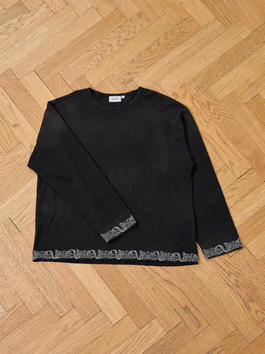 The FOUND Metallic Long Sleeve Embroidered Tee features a subtle patterned trim on the hem and cuffs, a unisex relaxed fit, and is shown laid flat on a wooden herringbone floor.