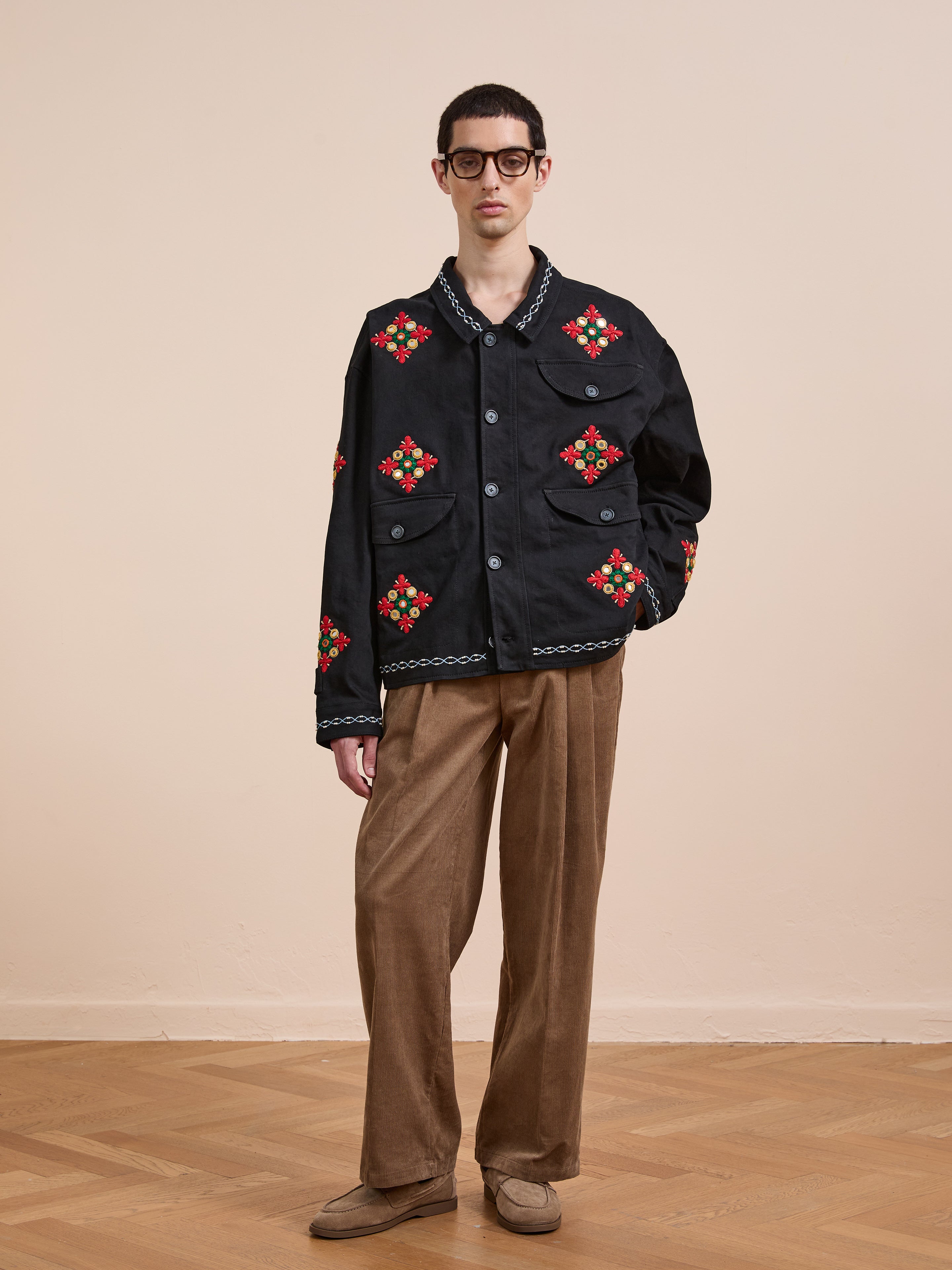 Indoors, a person wears Found's Mirror Ornament Hand Embroidered Work Jacket with red floral details, brown corduroy pants, glasses, and brown shoes against a beige background.