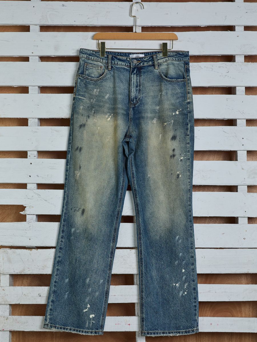 The Monet Paint Jeans by FOUND, a unisex relaxed fit featuring vintage blue wash with faded patches and paint splatters, hang on a wooden hanger against a white wooden backdrop.