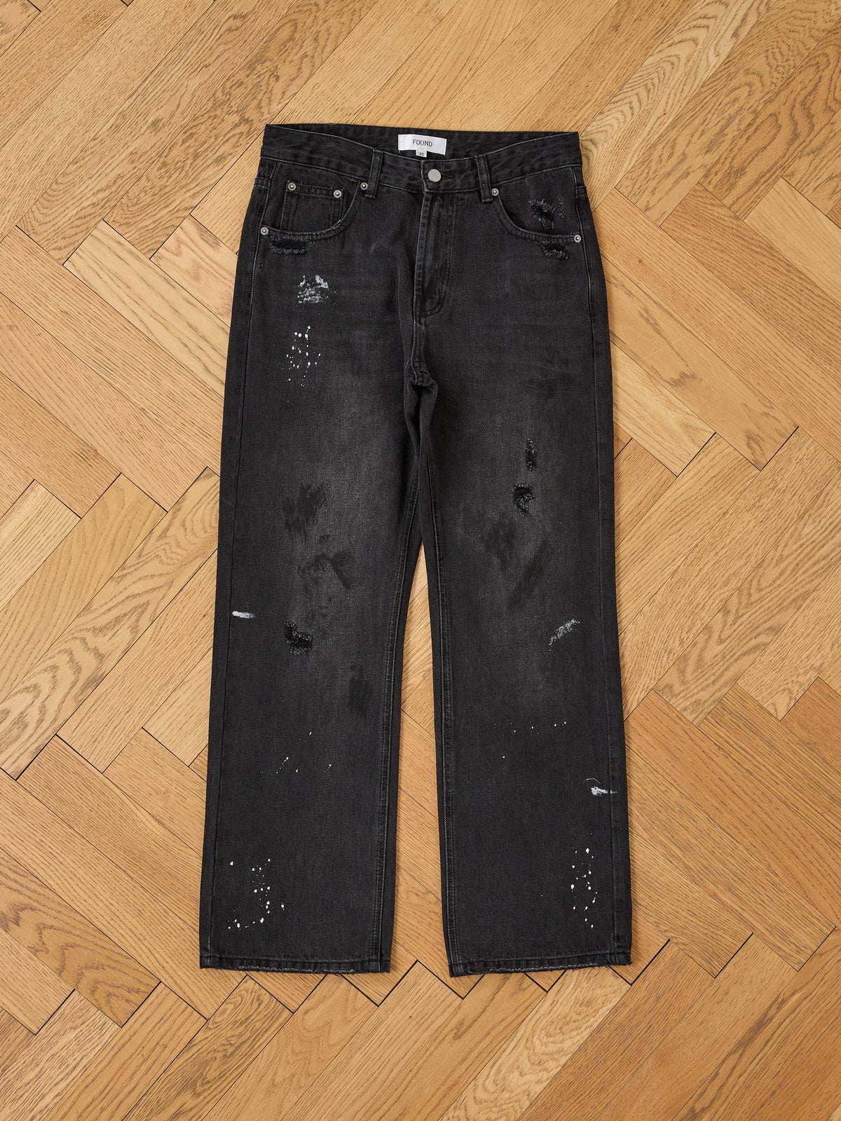 Monet Paint Jeans by FOUND, a vintage black denim with straight leg and distressed details, are laid flat on a light wood herringbone floor.